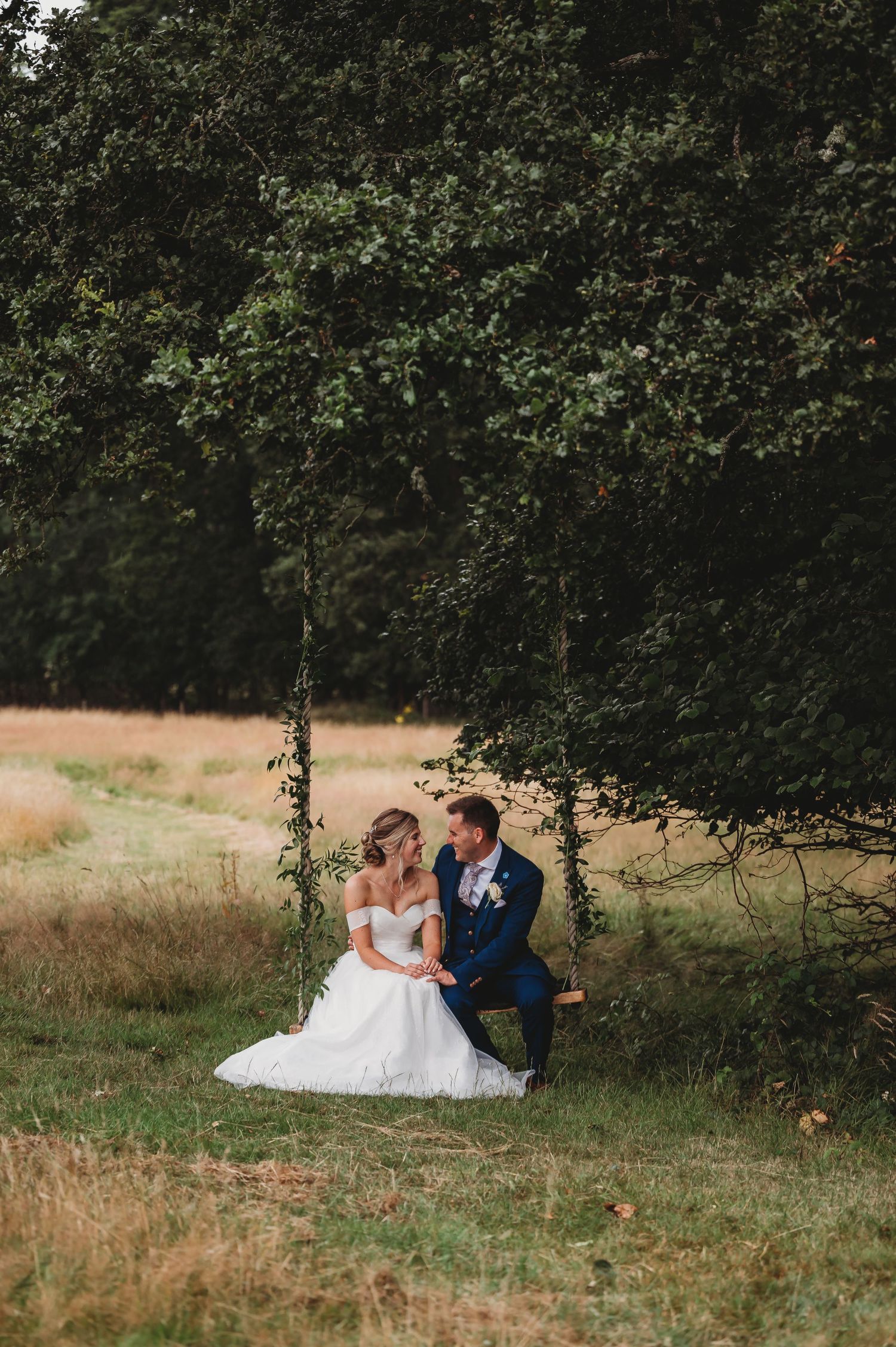 A couple dressed in wedding attire sits together on a rustic wooden swing beneath leafy green trees in a meadow.