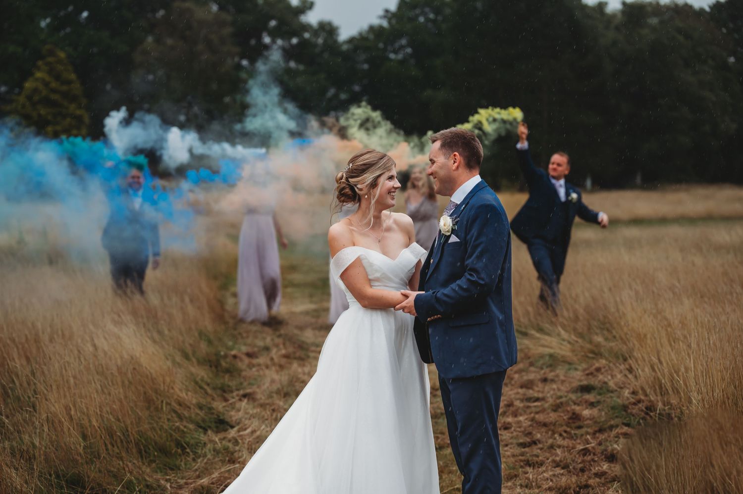 Wedding couple embraces in a golden field while blue smoke creates a dreamy atmosphere in the background.