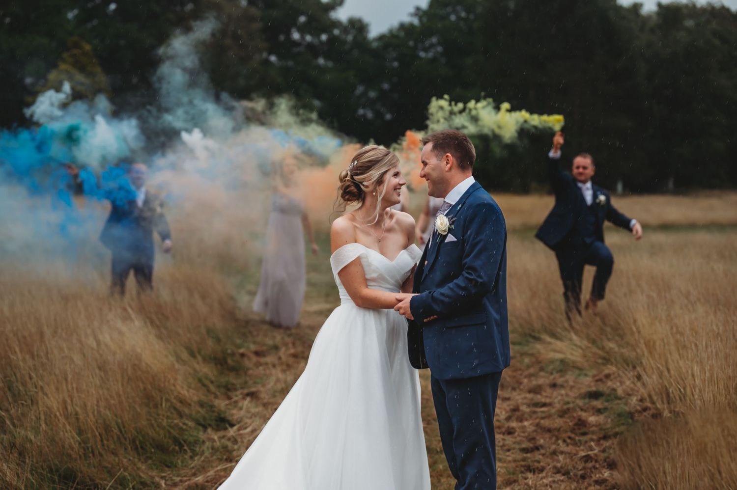 Newlyweds embrace in a field while colorful smoke bombs create a dramatic backdrop.