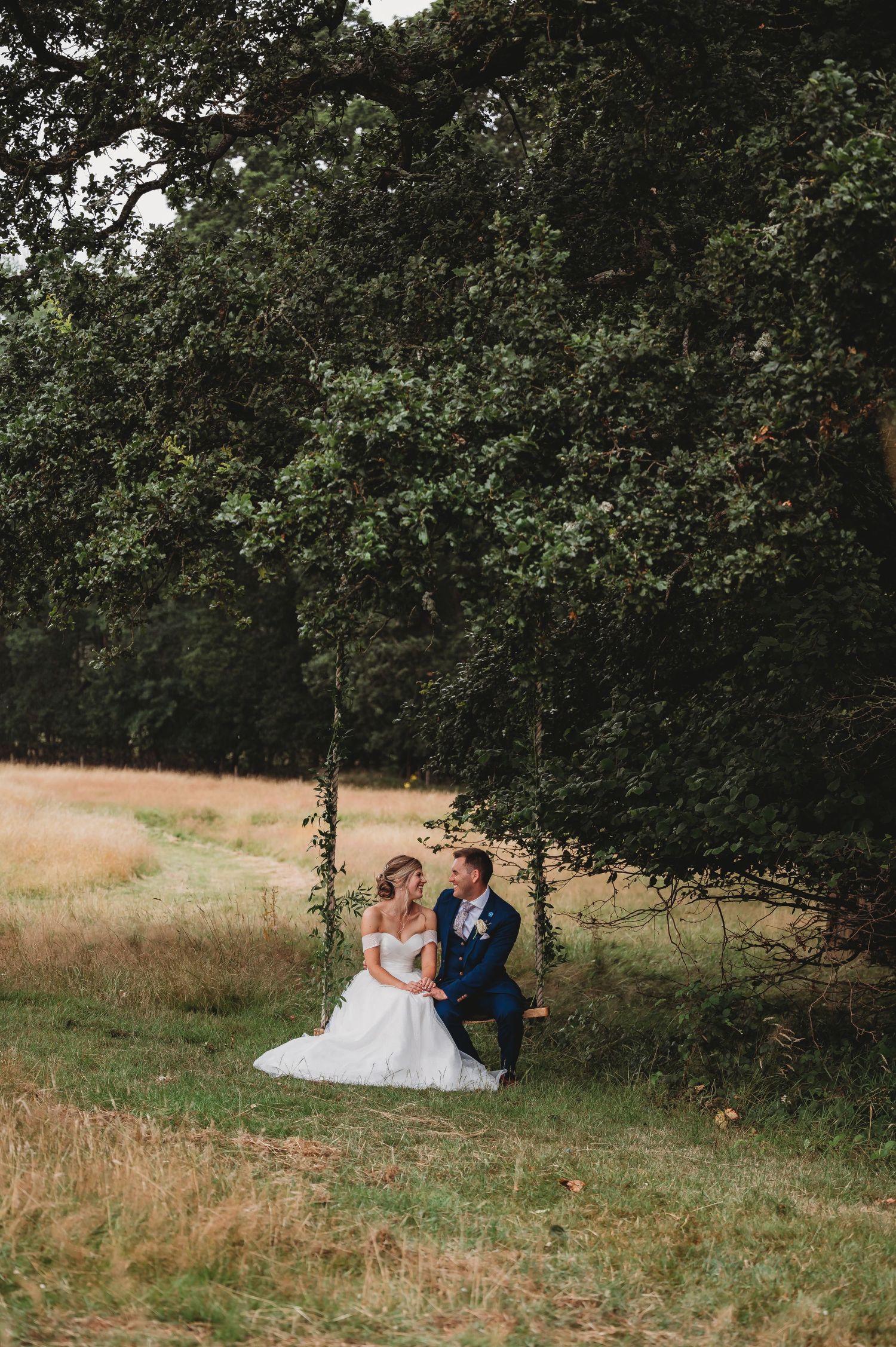 A couple sits together on a rustic wooden swing under a canopy of trees in a peaceful meadow setting.