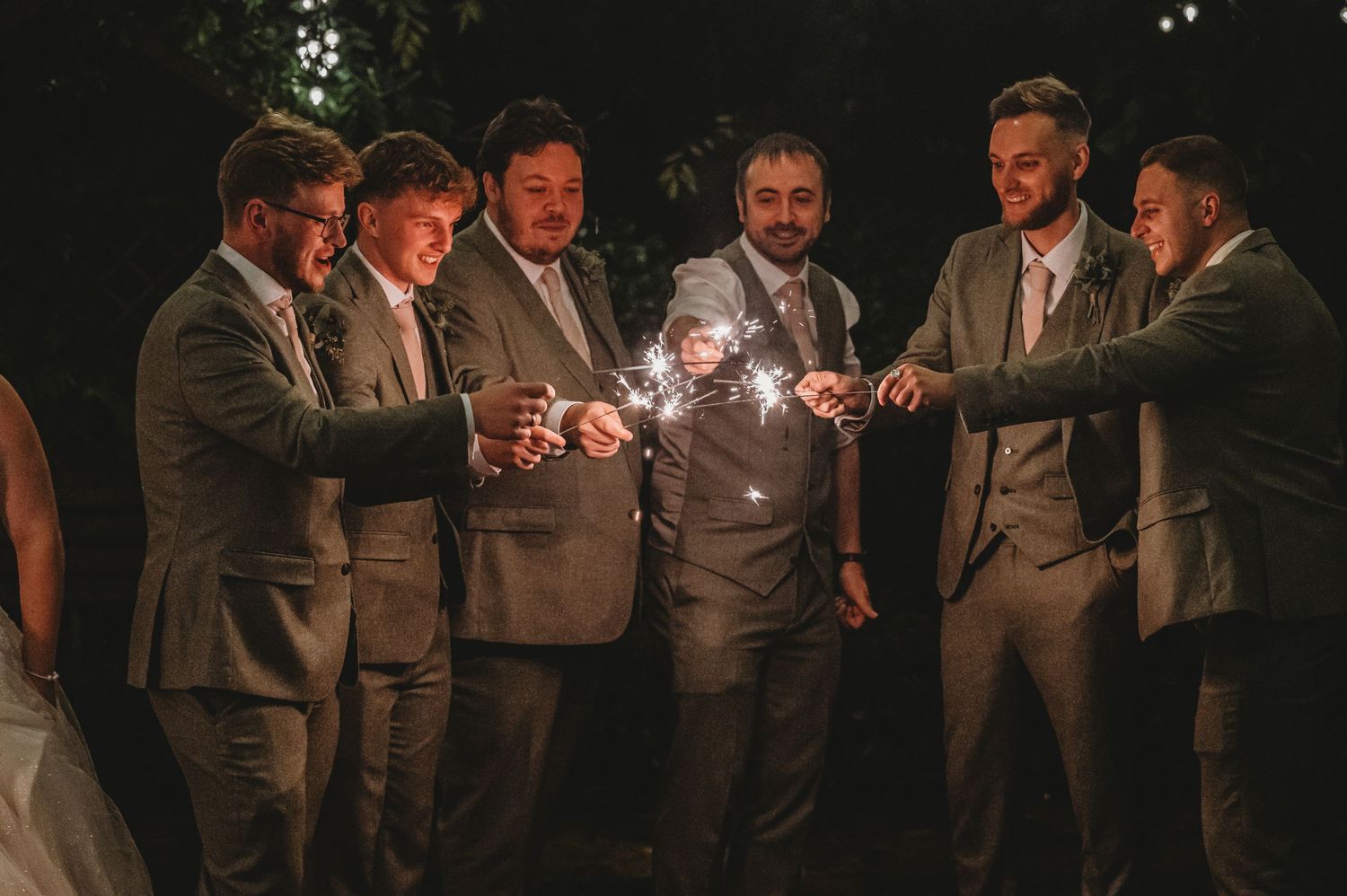 Groomsmen in gray suits gather with sparklers during evening wedding celebration.