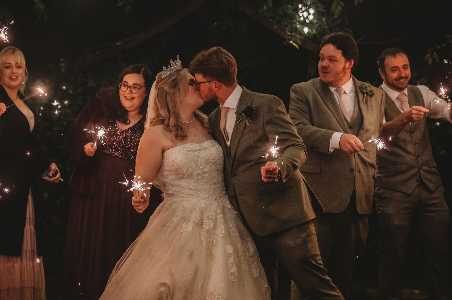 Wedding party holds sparklers during nighttime celebration outdoors.