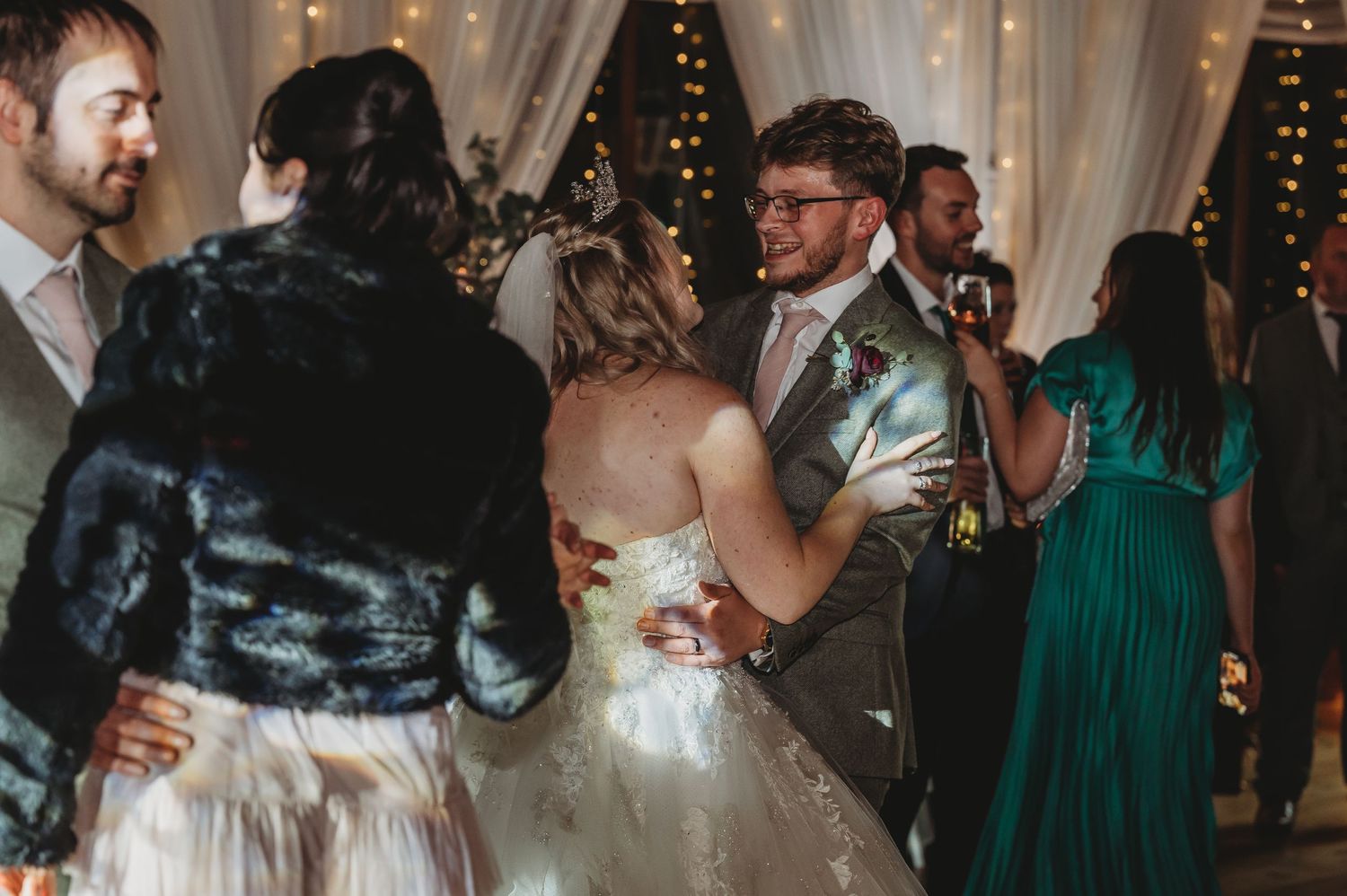 Newlyweds dancing among guests at their wedding reception with decorative lighting and white drapes.