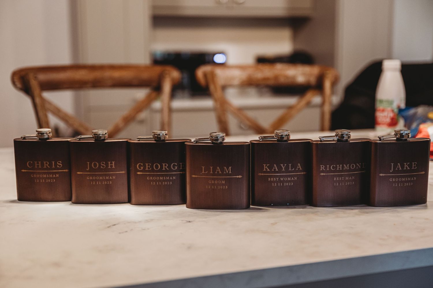 Row of dark brown personalized flasks lined up on a beige surface with wooden chairs in the background.