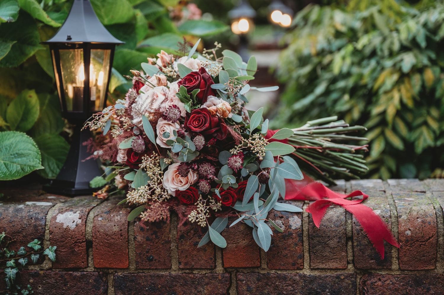 A romantic wedding bouquet with pink and red flowers rests on a brick wall next to a garden lantern.