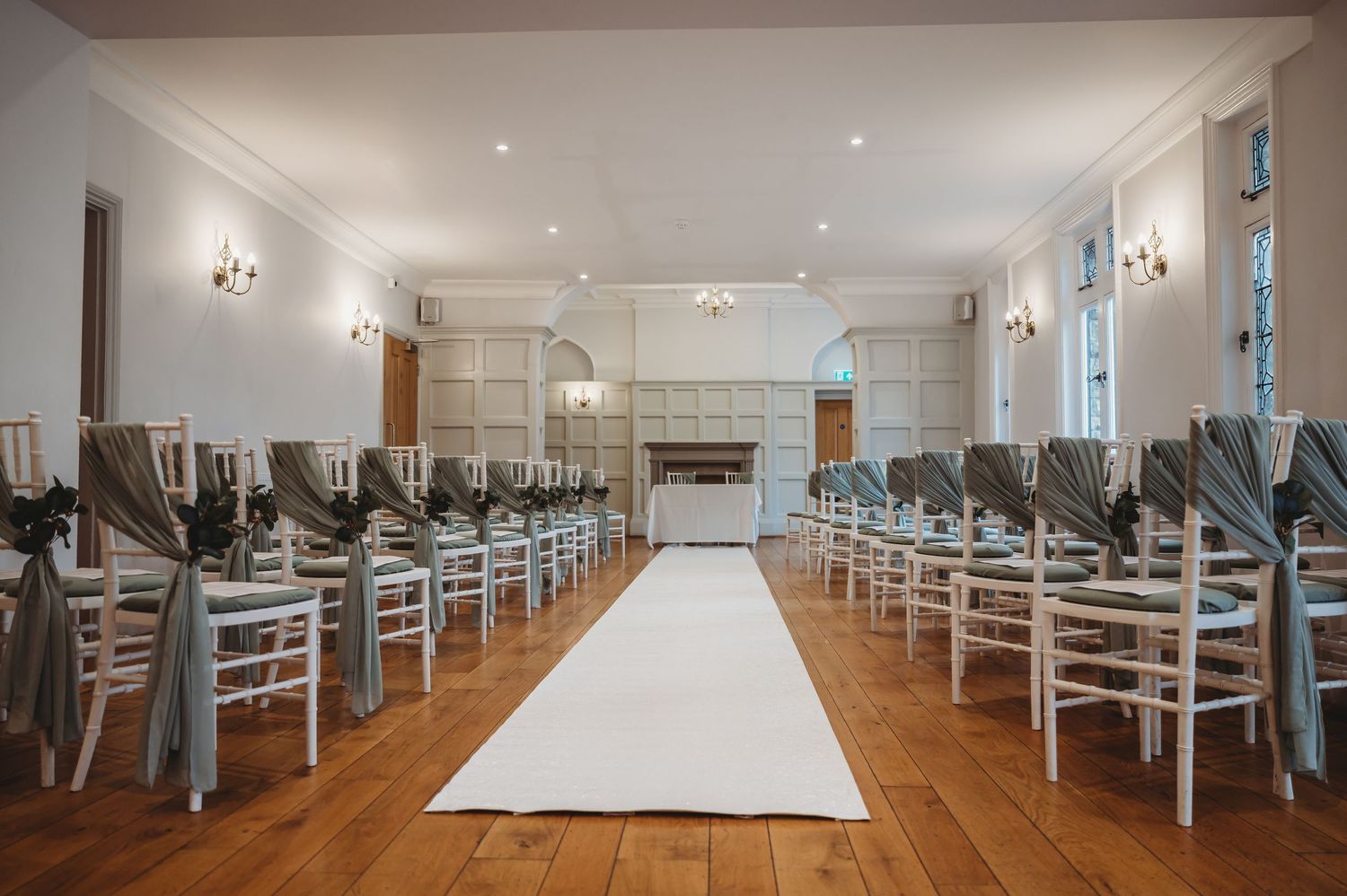 White chairs line both sides of an aisle runner in an elegant wedding ceremony venue with hardwood floors.