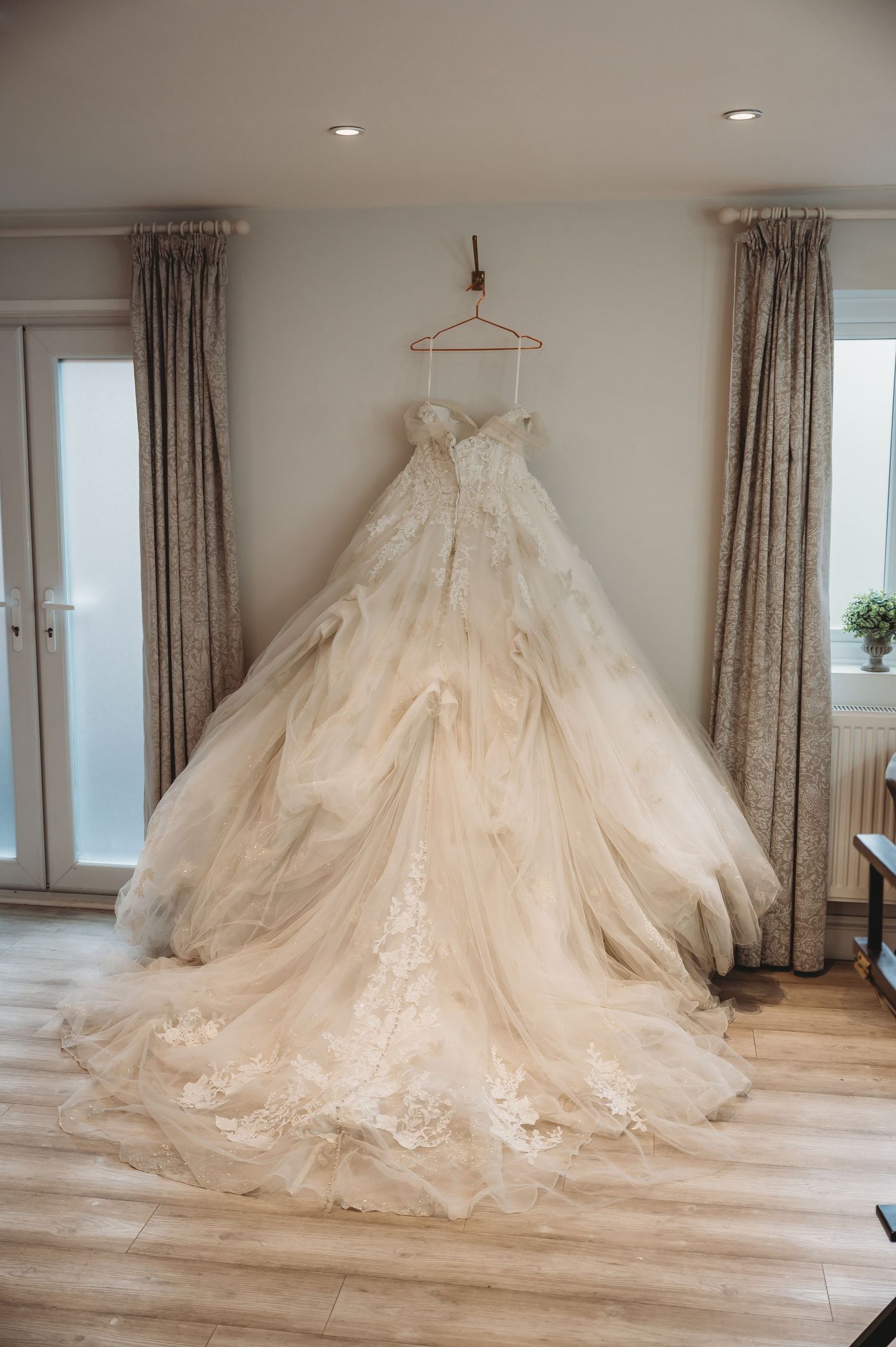 A luxurious white lace wedding dress with a dramatic tulle train hangs on display against neutral curtains.