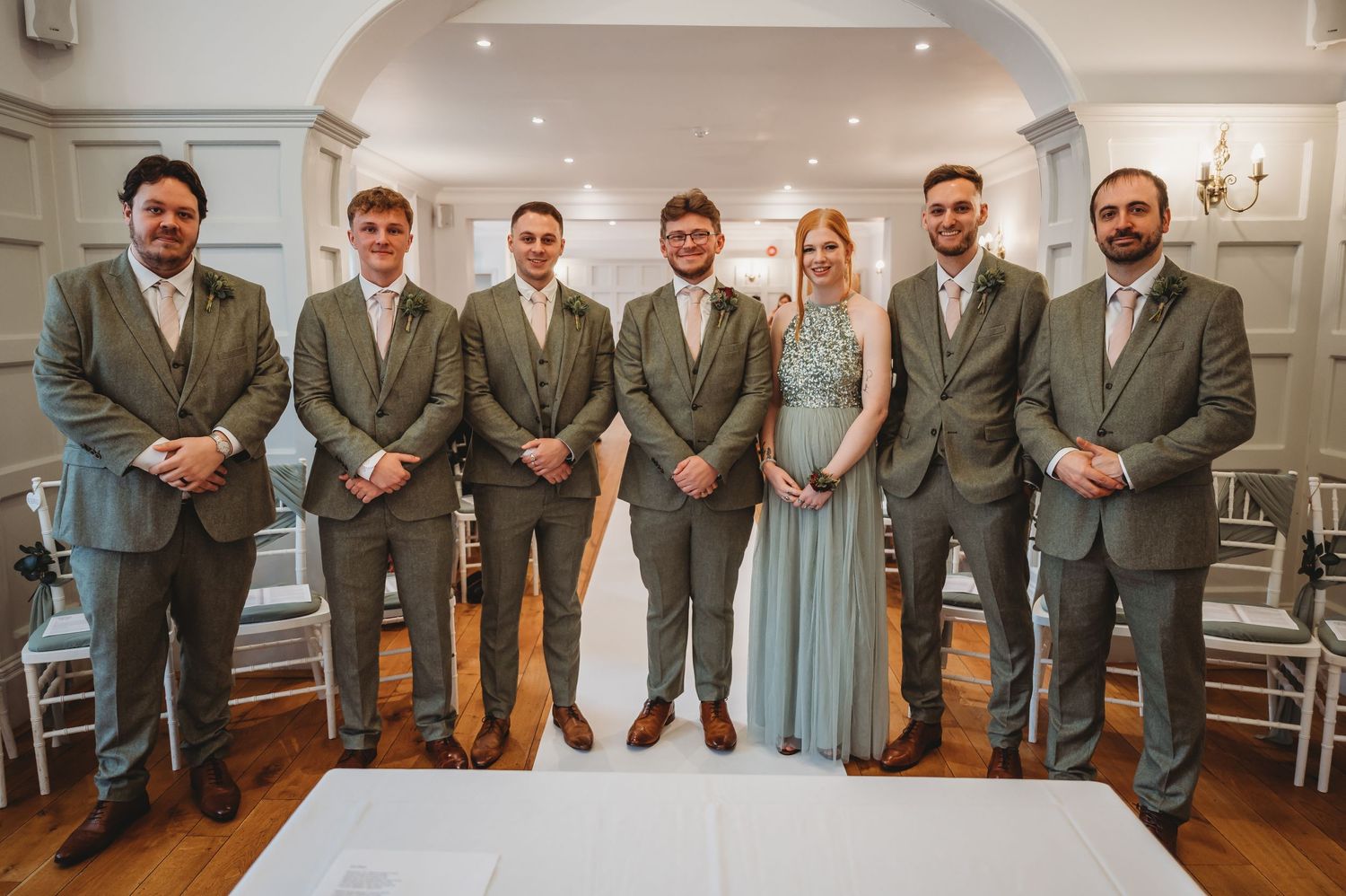 Wedding party members in coordinated gray suits pose formally in an elegant white room.