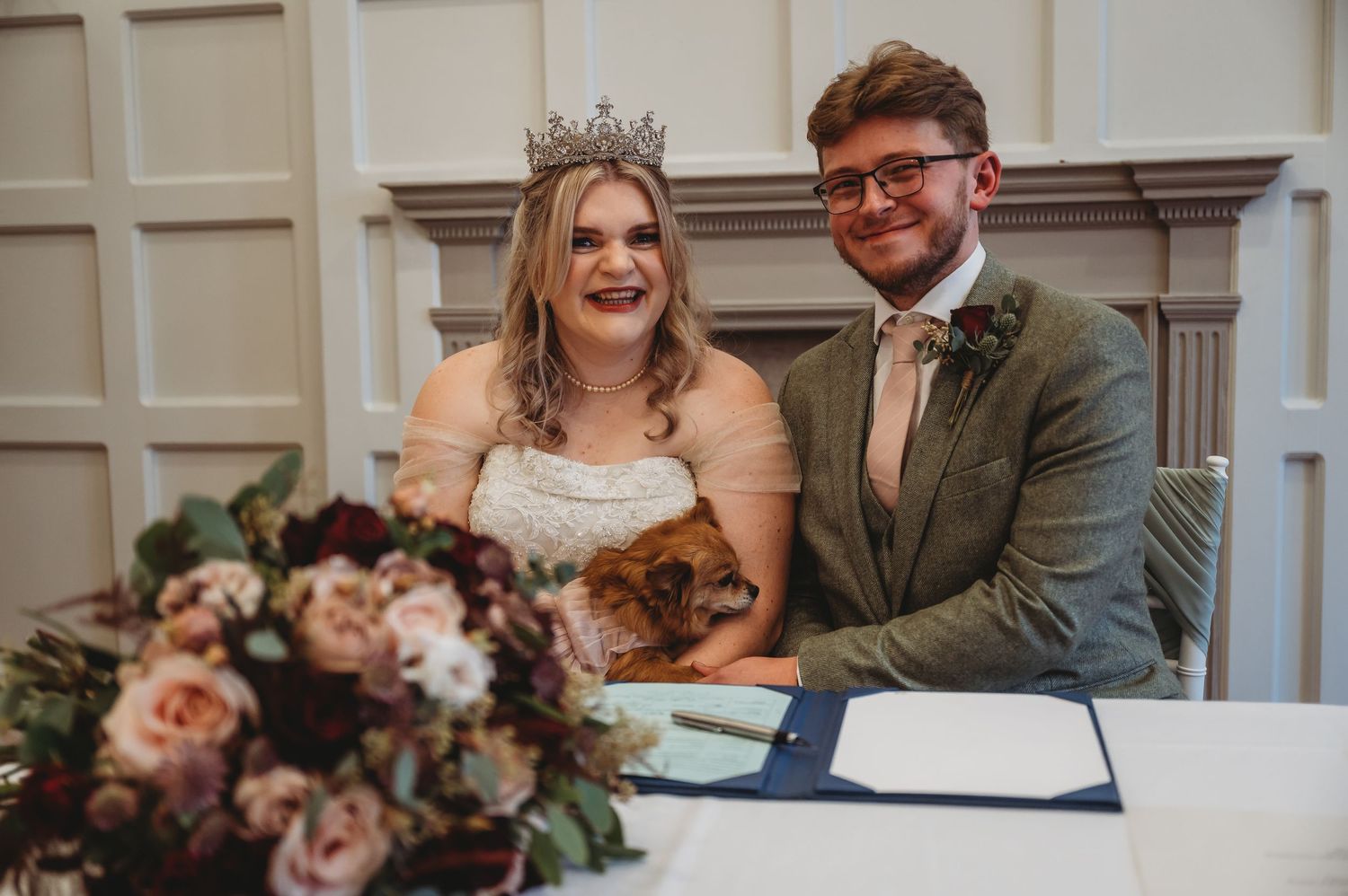 Newlyweds laugh together at a decorated signing table with pink floral arrangements.