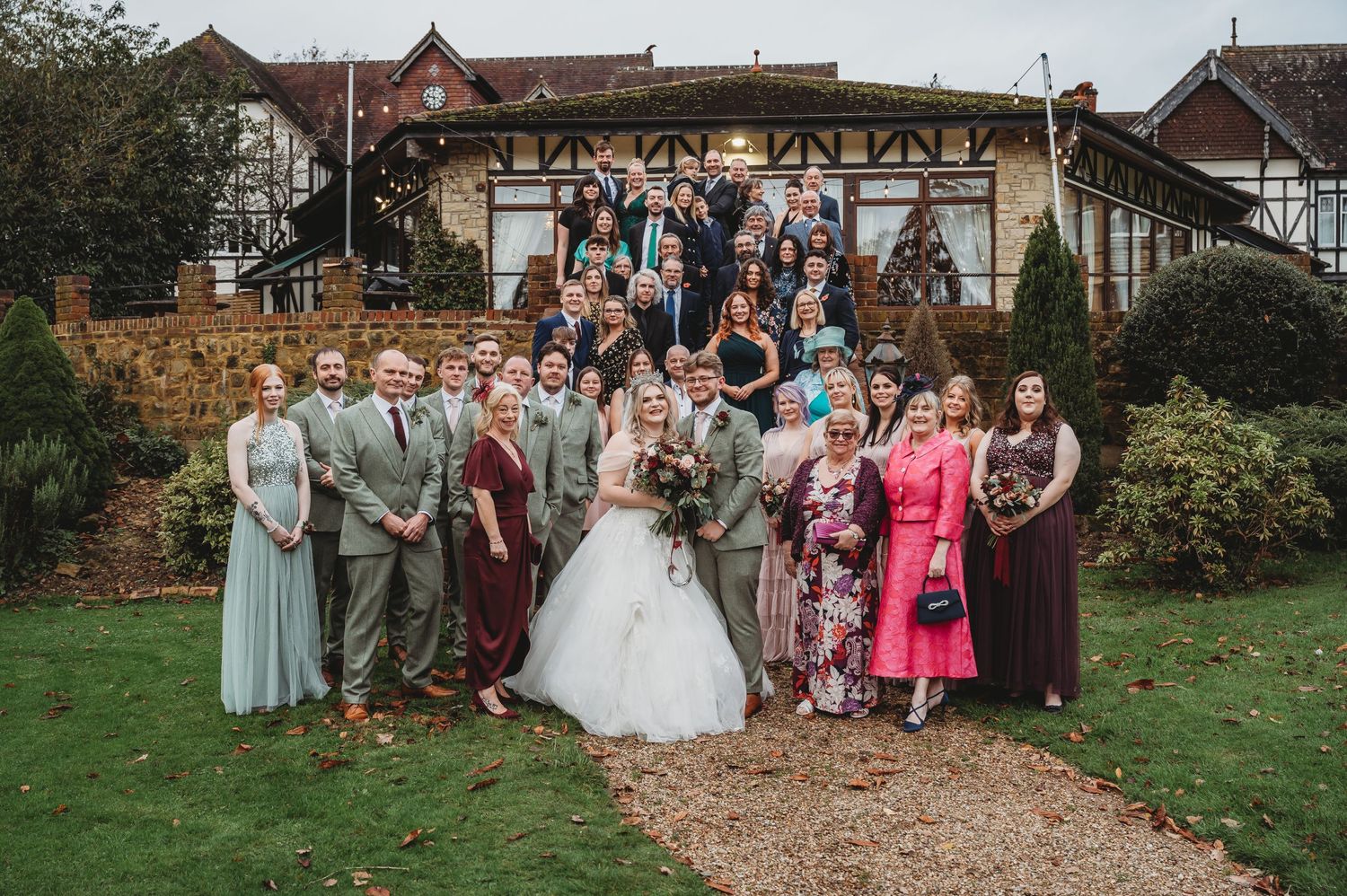 Large wedding party poses together outside a rustic venue on a grassy lawn with decorative stone walls.