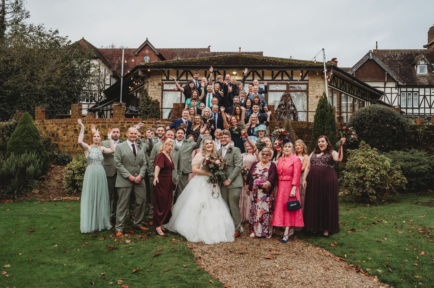 Large wedding party poses on outdoor garden steps of Tudor-style venue.