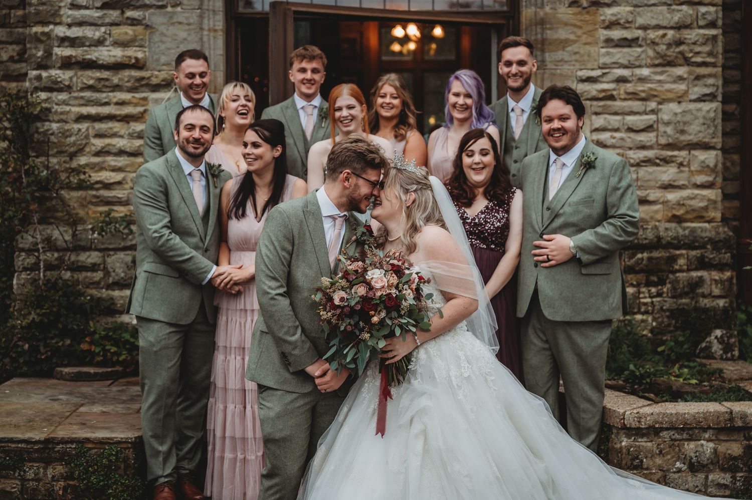Wedding party in sage green and pink attire posed formally against stone wall.