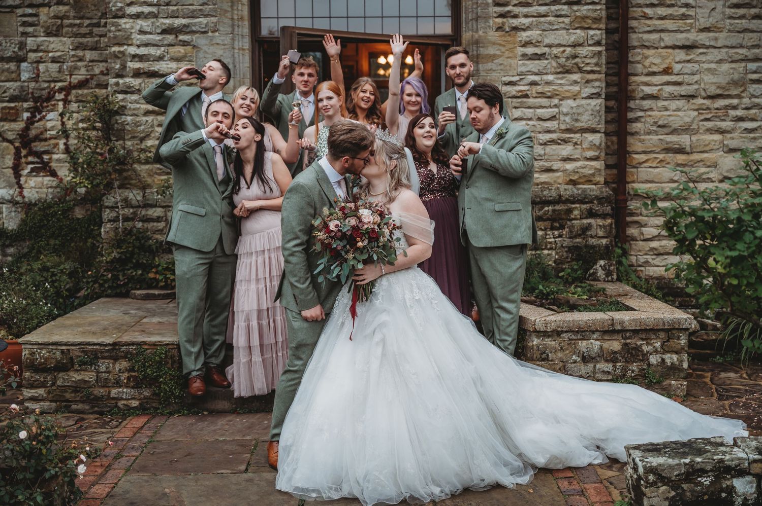 Festive wedding party celebrates with silly expressions outside historic building.