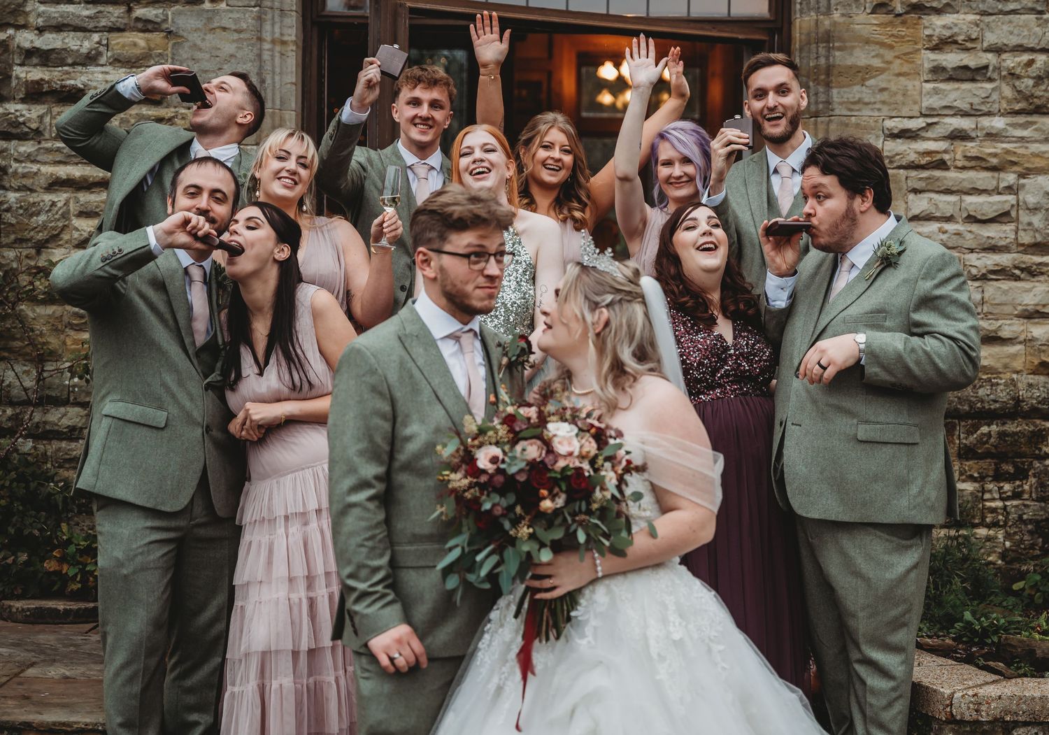 Wedding party strikes playful poses together against stone wall backdrop.