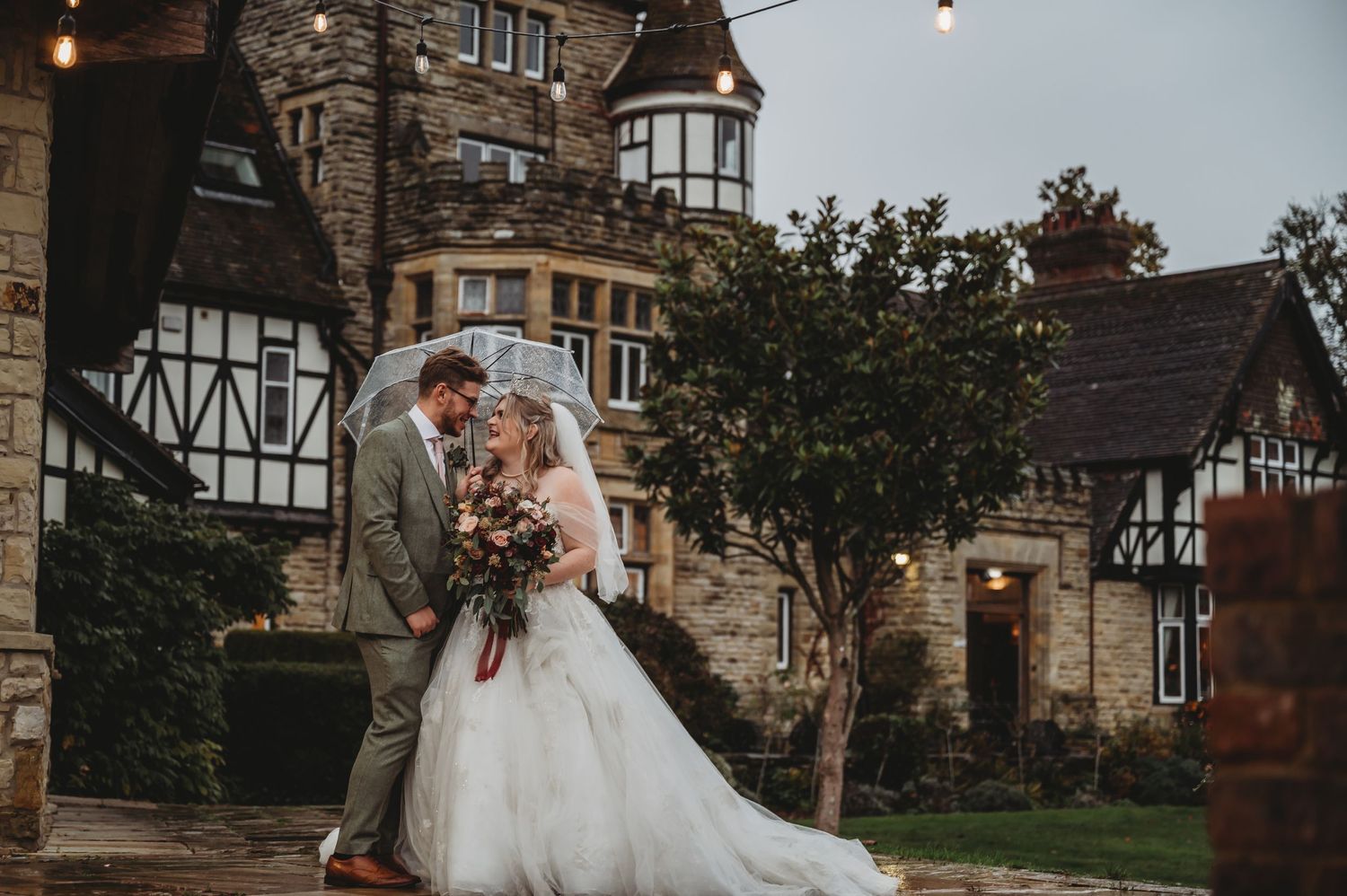 A romantic wedding couple poses outside a Tudor-style stone mansion on overcast day.