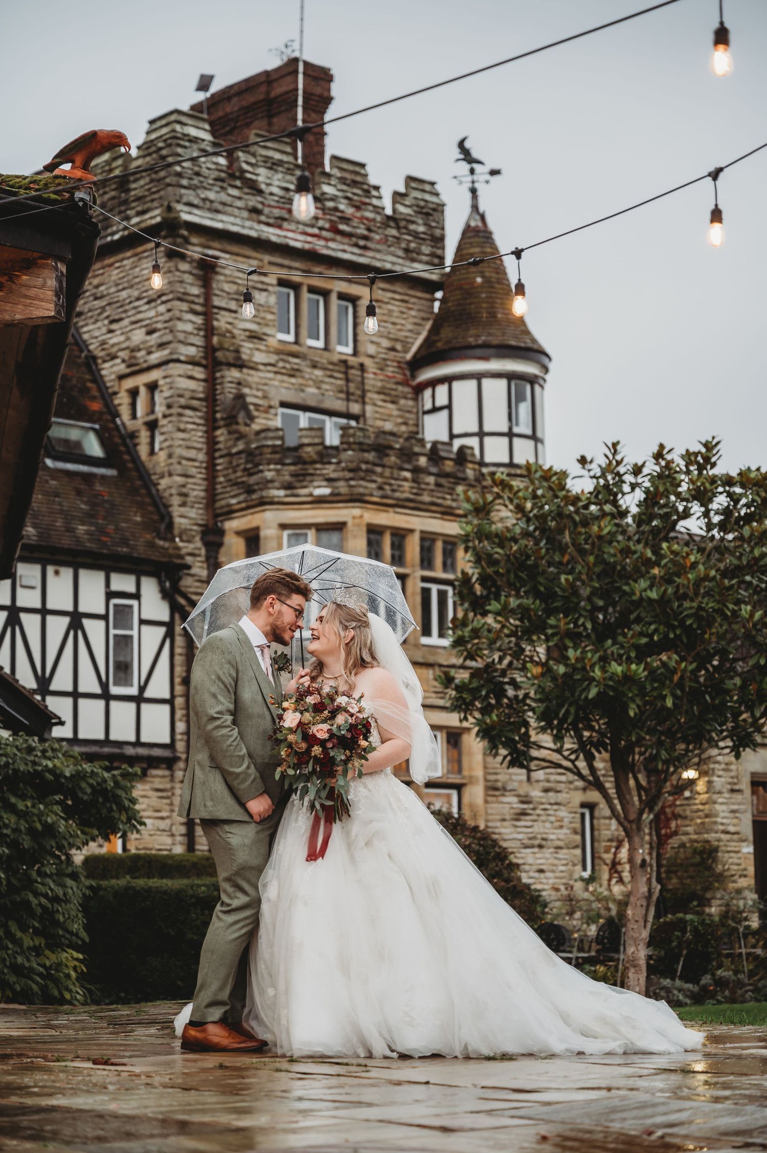 Couple shares intimate moment outside stone castle with string lights and Tudor architecture.