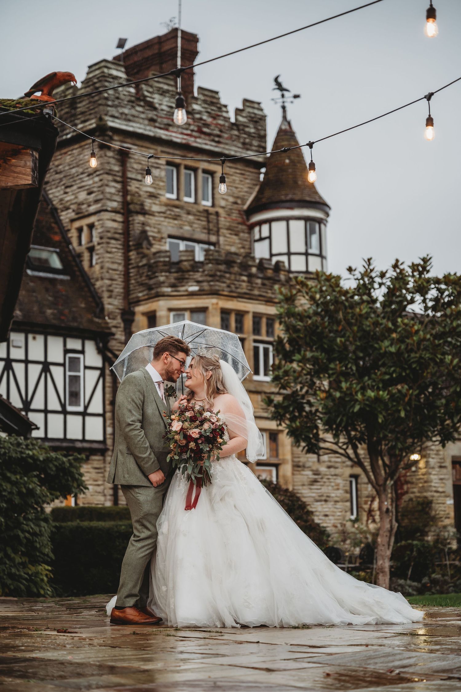 Rainy day wedding portrait outside historic stone castle with string lights and turret.