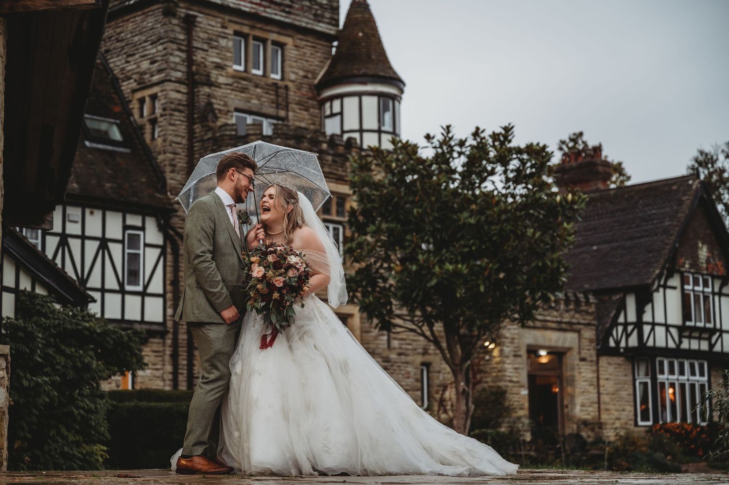 Wedding portrait in front of Tudor-style stone castle with turret on overcast day.