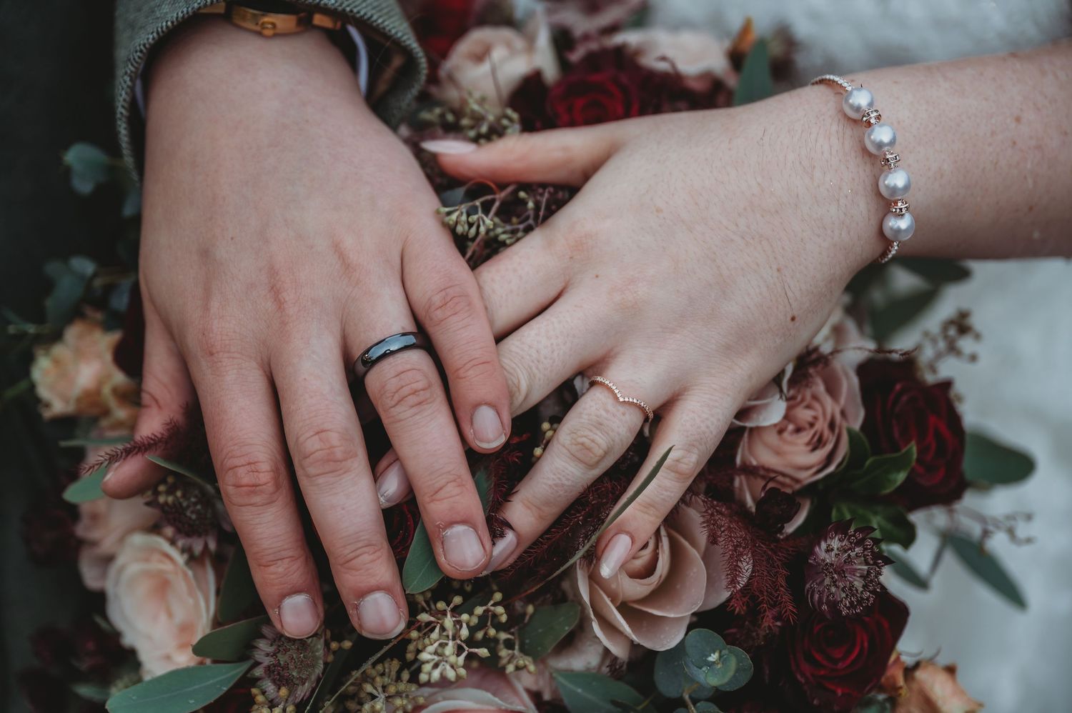 Close-up of wedding rings and hands resting on burgundy and blush rose bouquet.