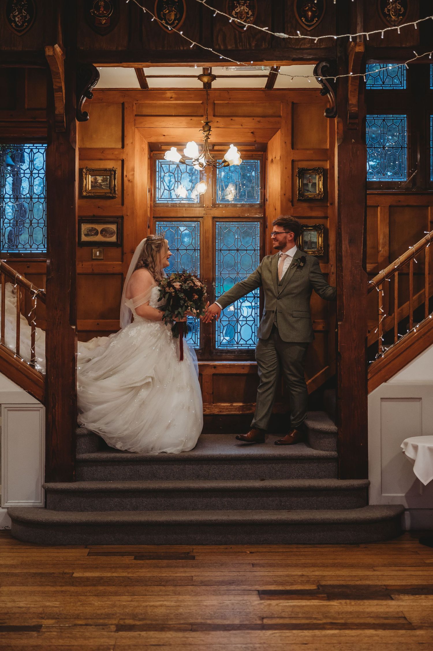 Wedding couple stands on wooden staircase in rustic venue with stained glass windows and string lights.