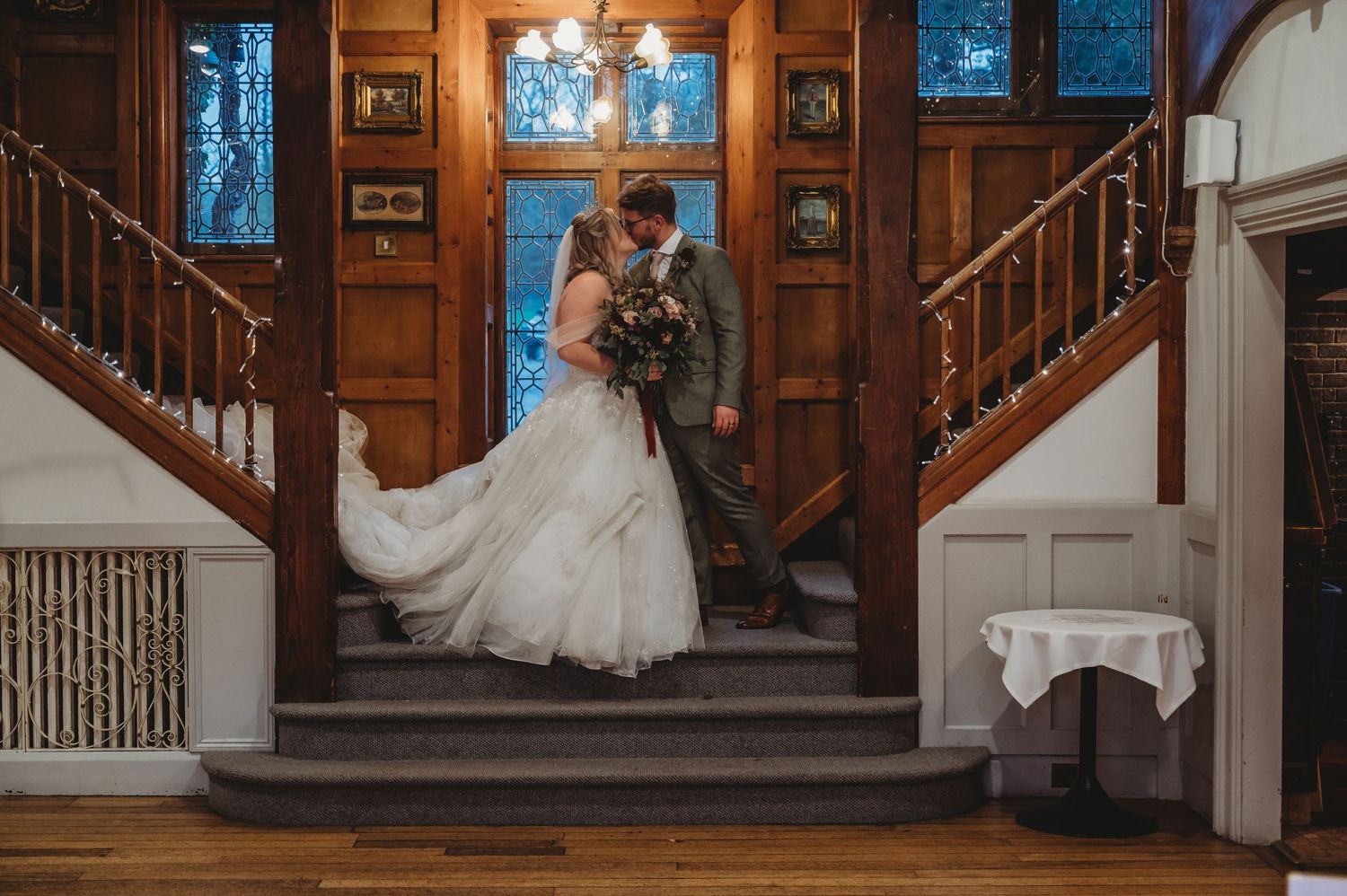 Romantic portrait of newlyweds on wooden staircase with stained glass windows in background.