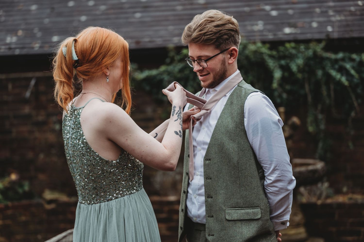 Someone in a sparkly green dress adjusts another person's tie outdoors.