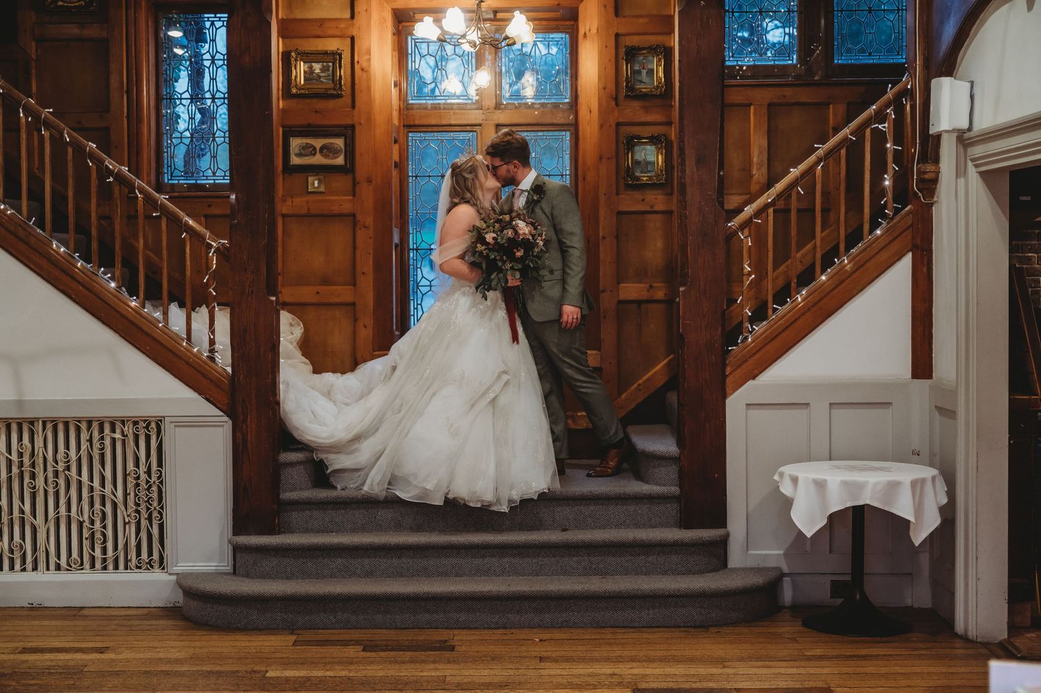 A wedding couple poses on wooden stairs in front of stained glass windows in an elegant venue.