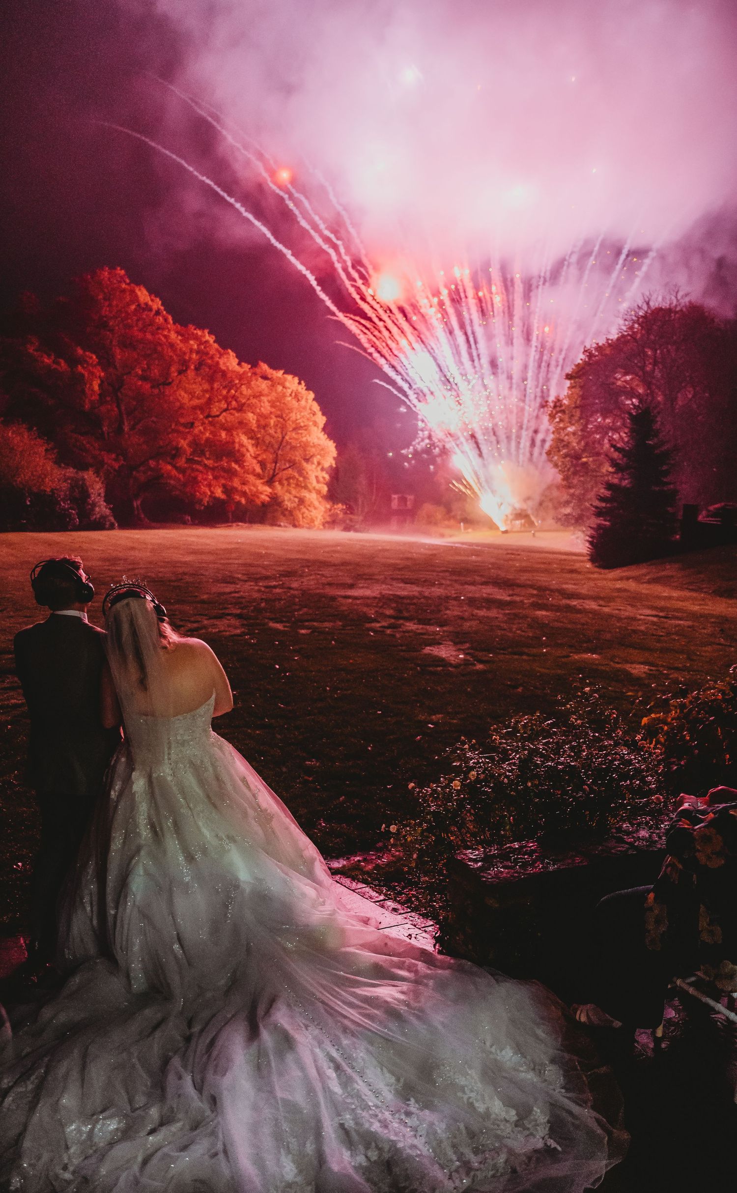Silhouetted couple watches vibrant red and pink fireworks display at their wedding reception.