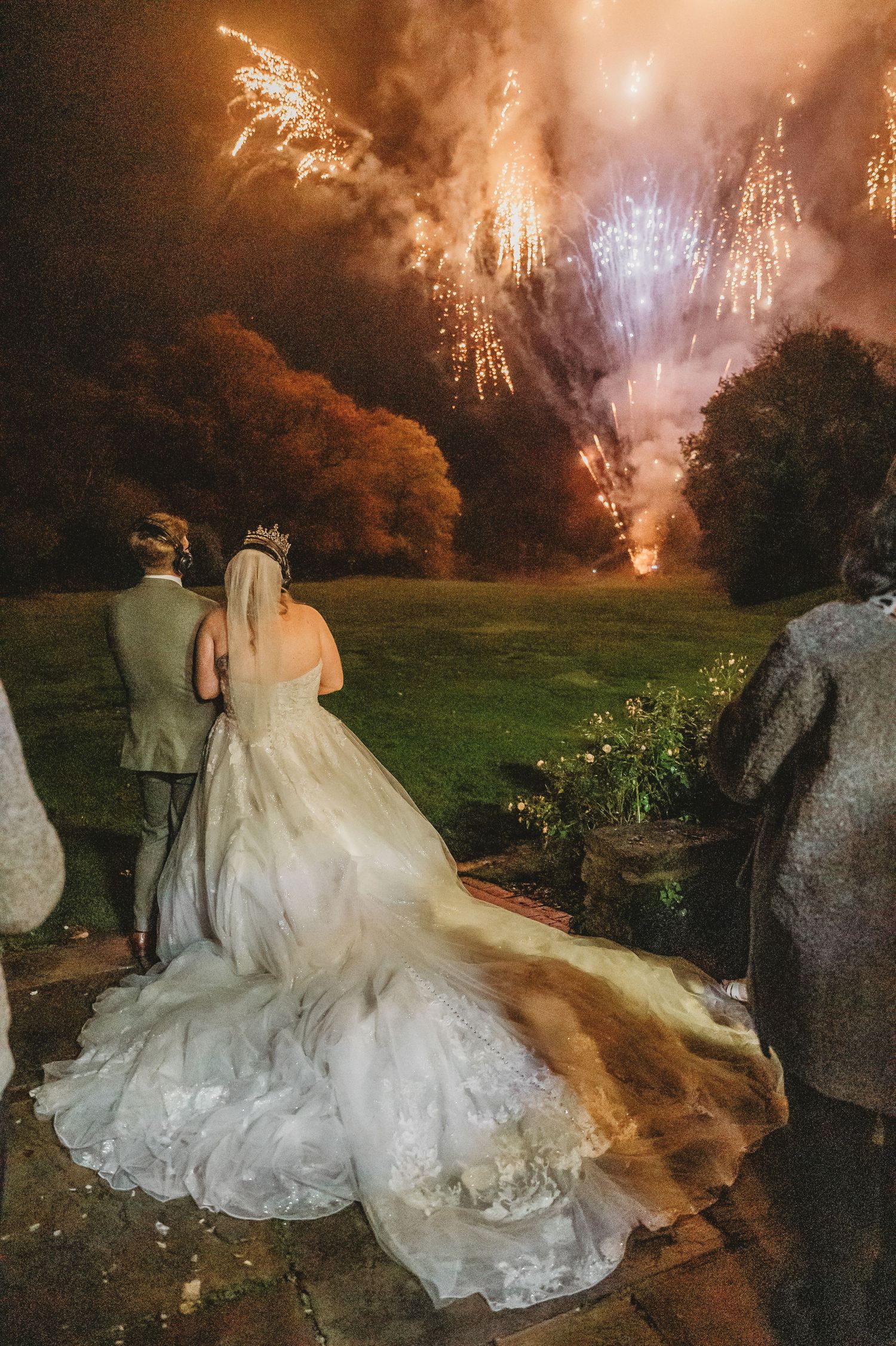 A bride in flowing white gown watches fireworks with her partner during outdoor wedding celebration.