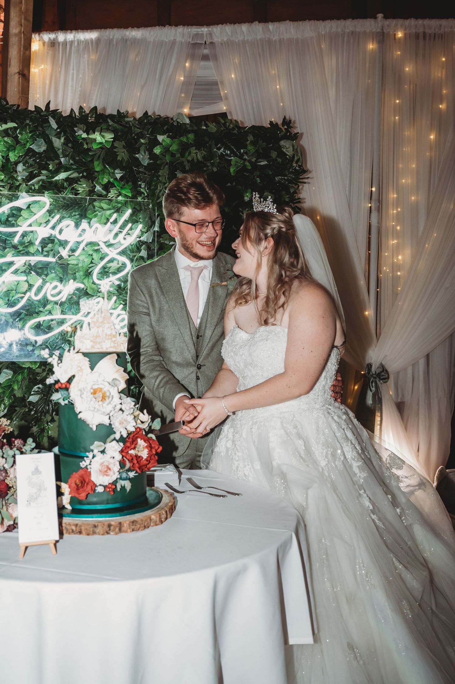 A couple cuts their elegant green and white wedding cake beside a floral backdrop with fairy lights.
