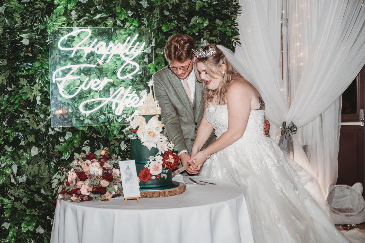 Newlyweds cut their wedding cake together in front of a green hedge wall with neon signage.