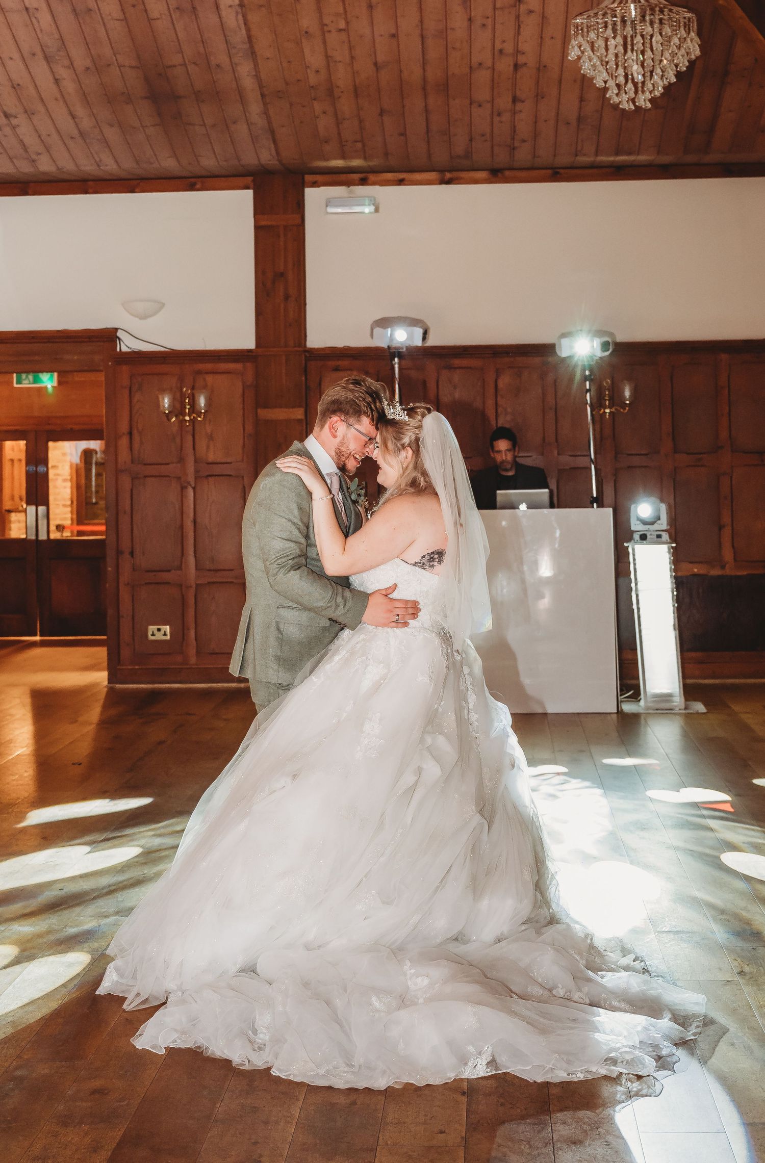 A couple shares their first dance in a wood-paneled reception hall with a flowing white wedding gown and sage green suit.