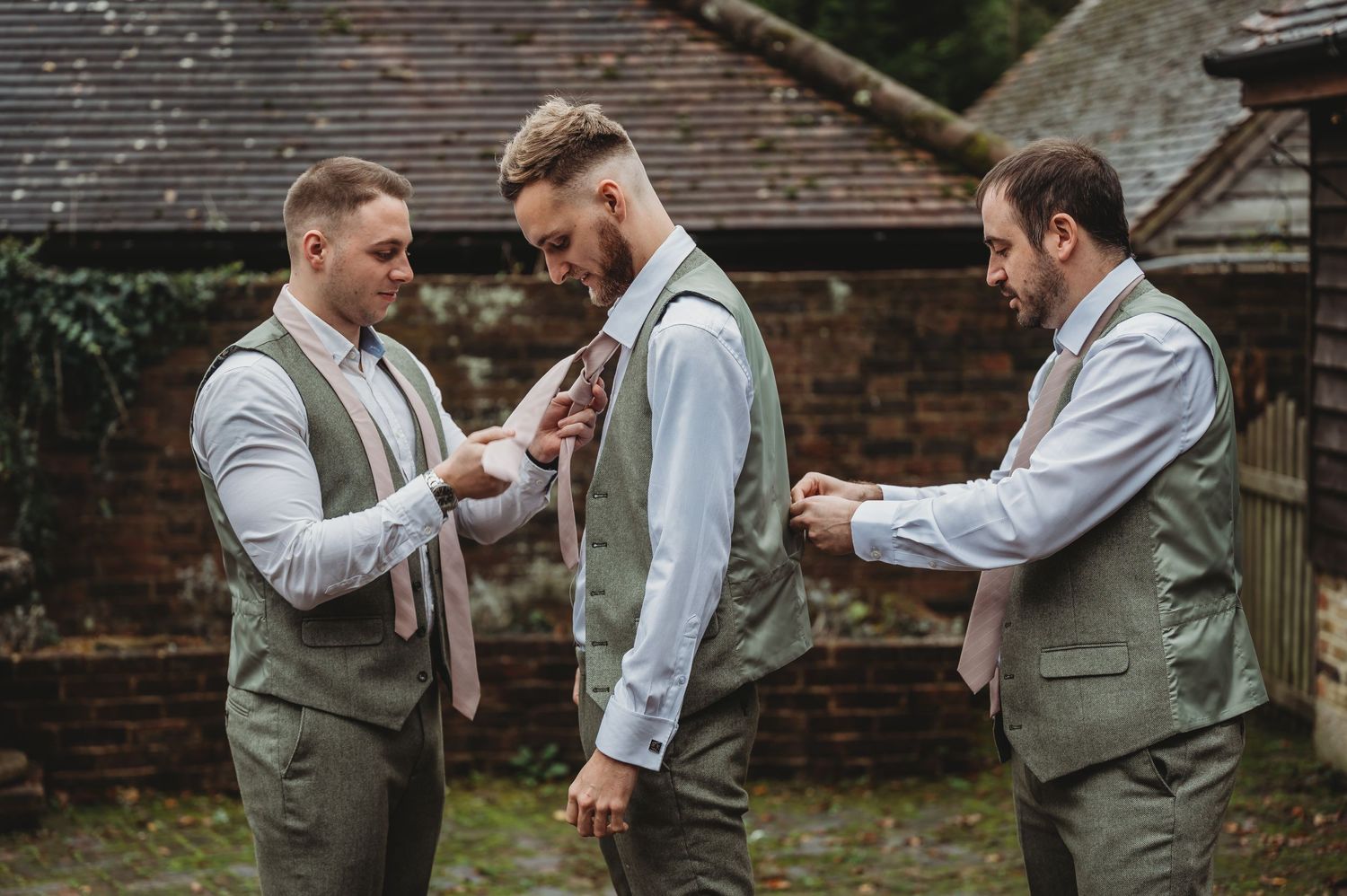 Three people in matching sage green vests help with formal wear preparations outdoors.