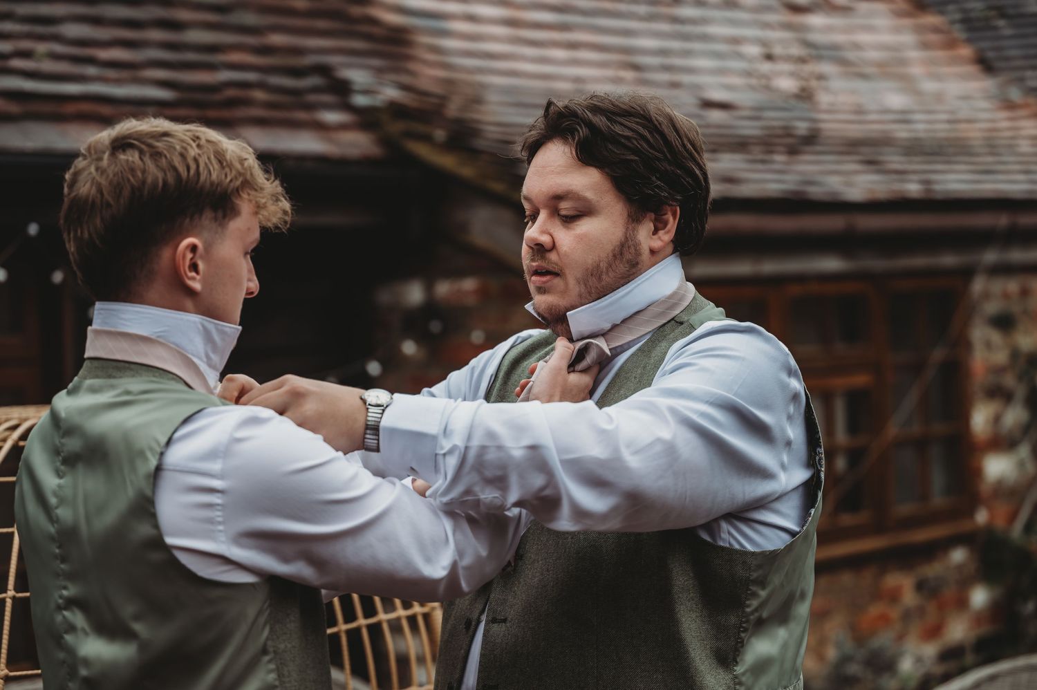 Two people help each other with formal wear adjustments against a brick building backdrop.