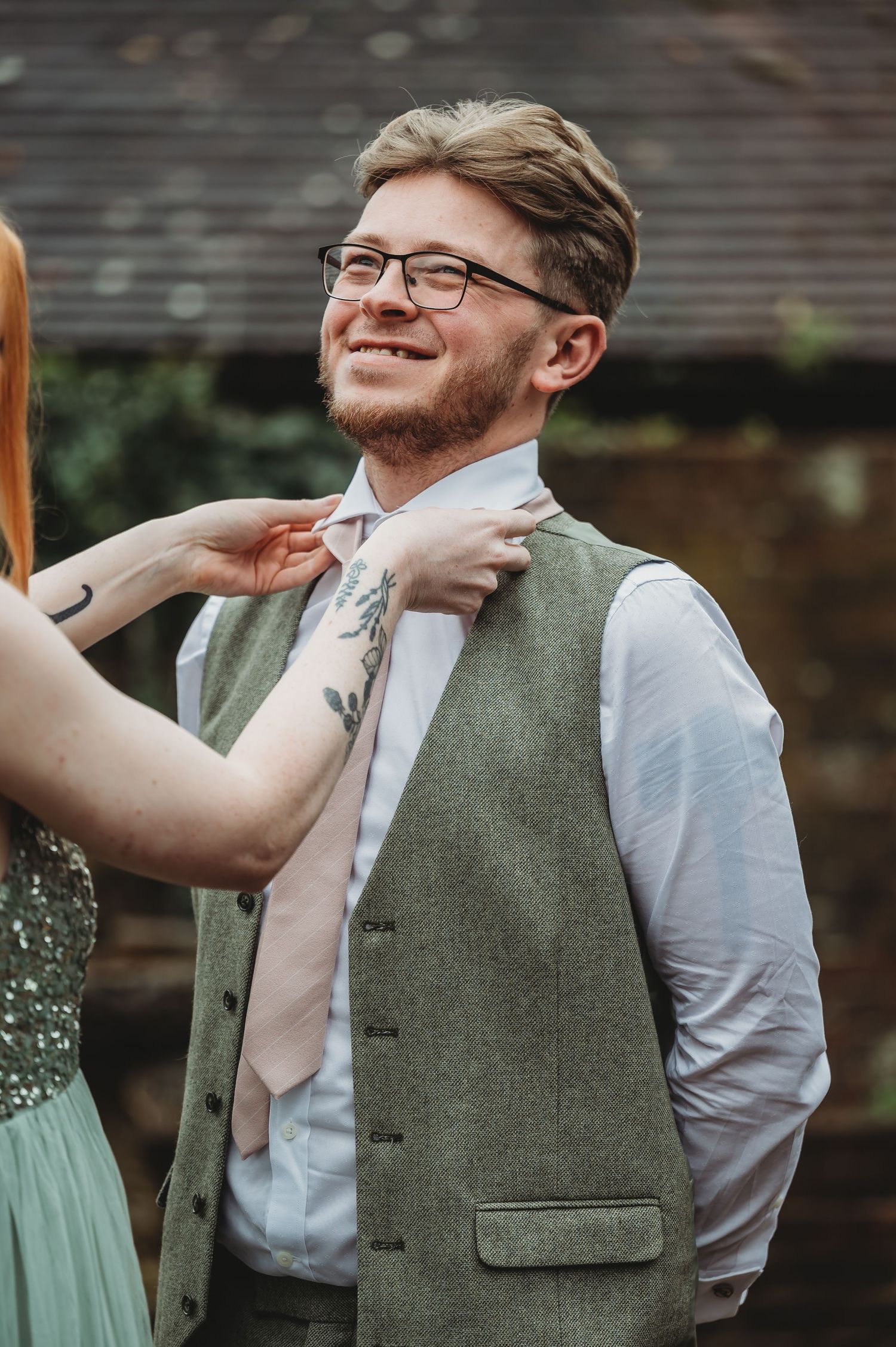 Someone helps adjust a pink tie on a person wearing a sage green vest.