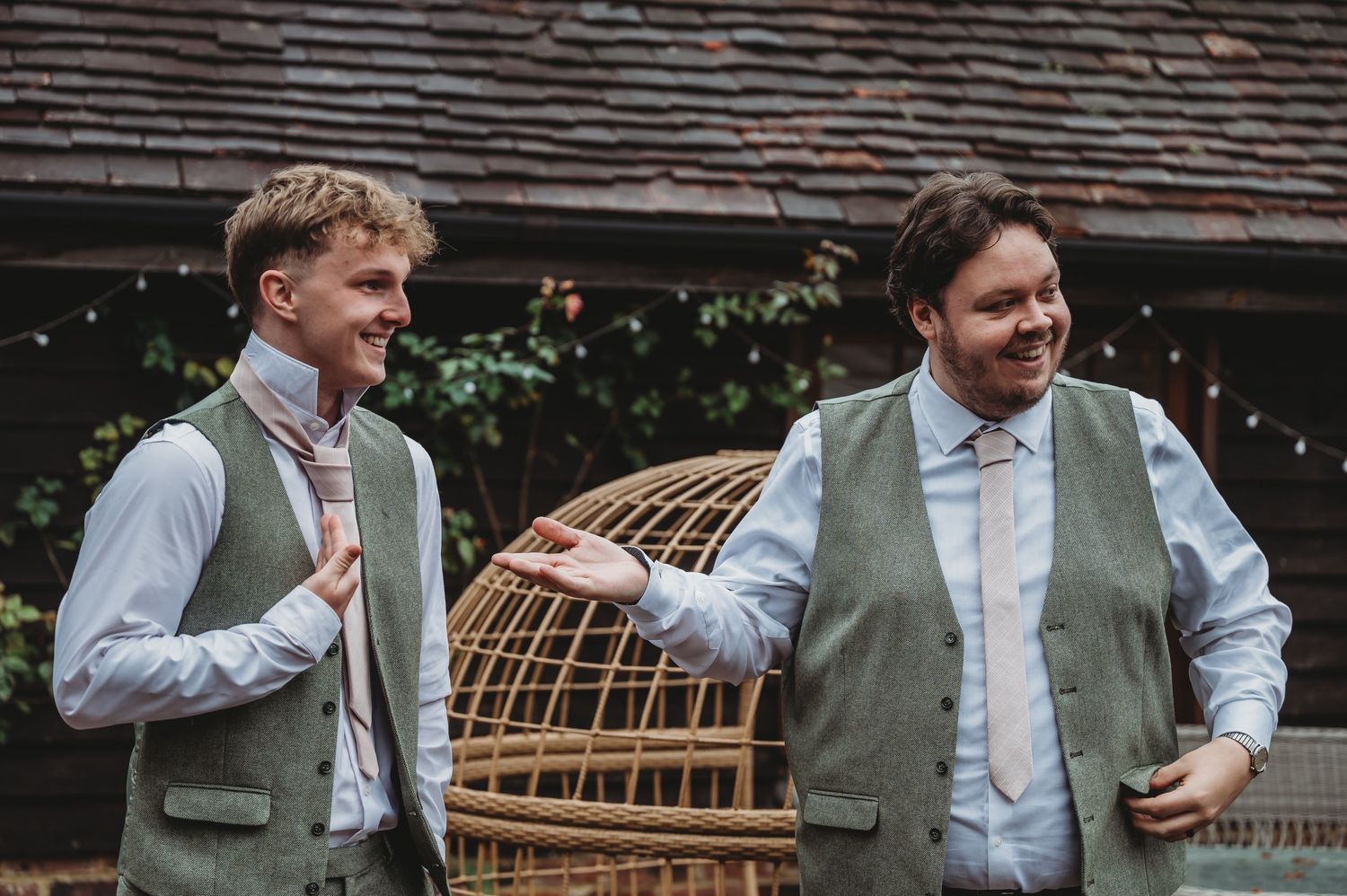 Two people in matching sage green vests stand chatting near a wicker structure outdoors.