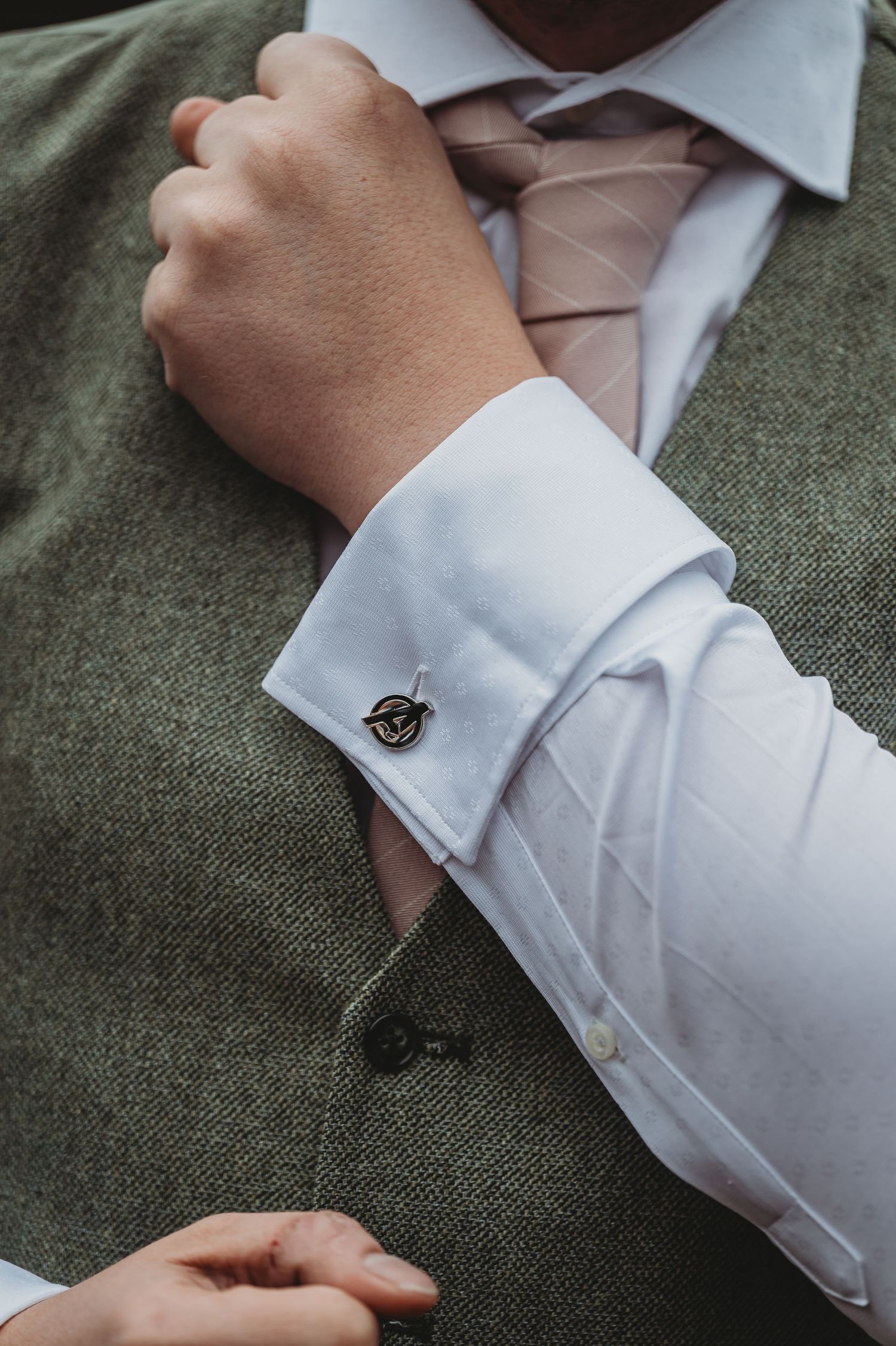 Close-up of a decorative cufflink on a white dress shirt sleeve with a pink tie and green suit jacket.