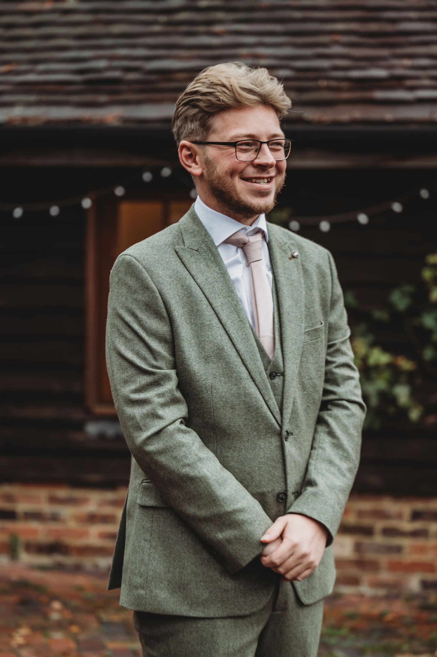 A well-dressed person in a sage green suit and pink tie smiles warmly at an outdoor venue.
