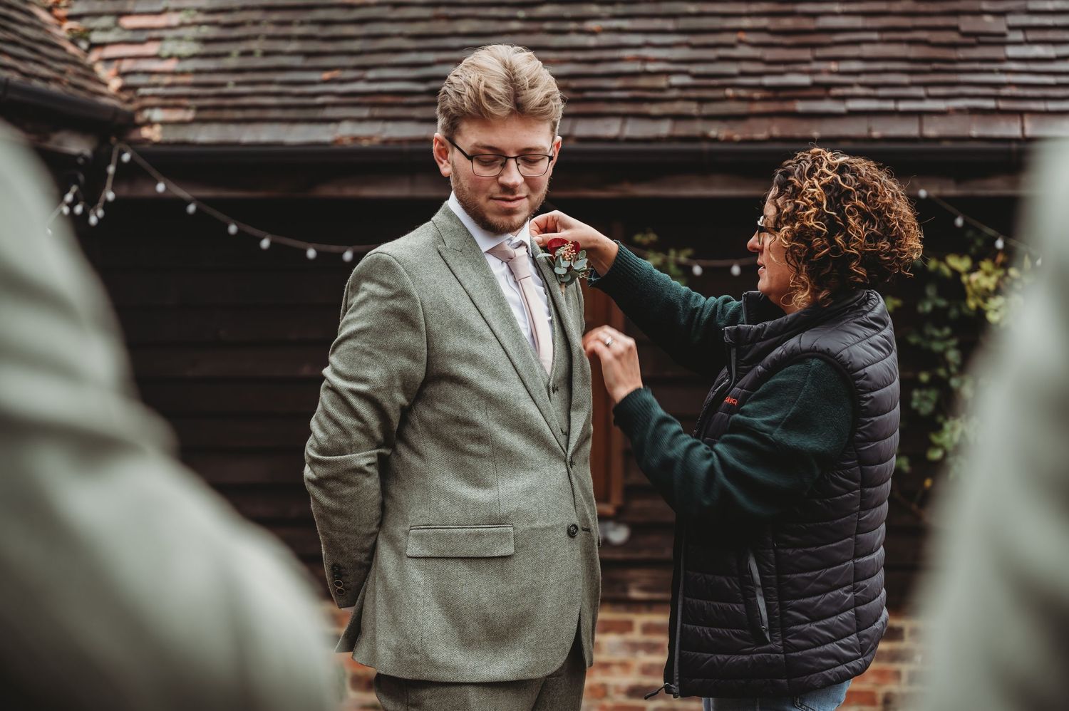 A stylist adjusts a boutonniere on a sage green suit jacket at a rustic venue.