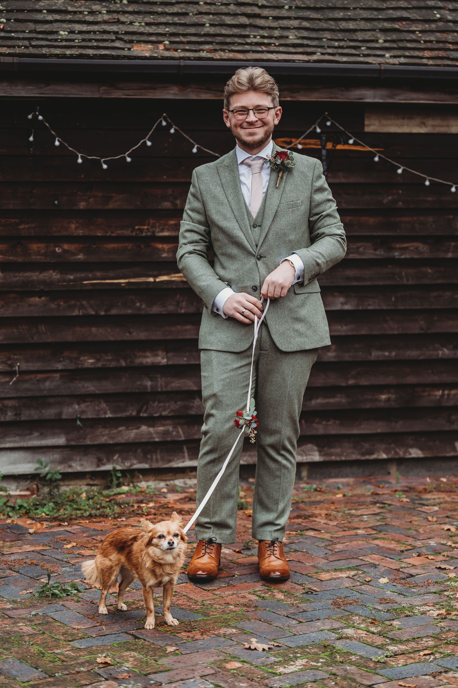 A small dog on a leash stands next to someone in a sage green three-piece suit.