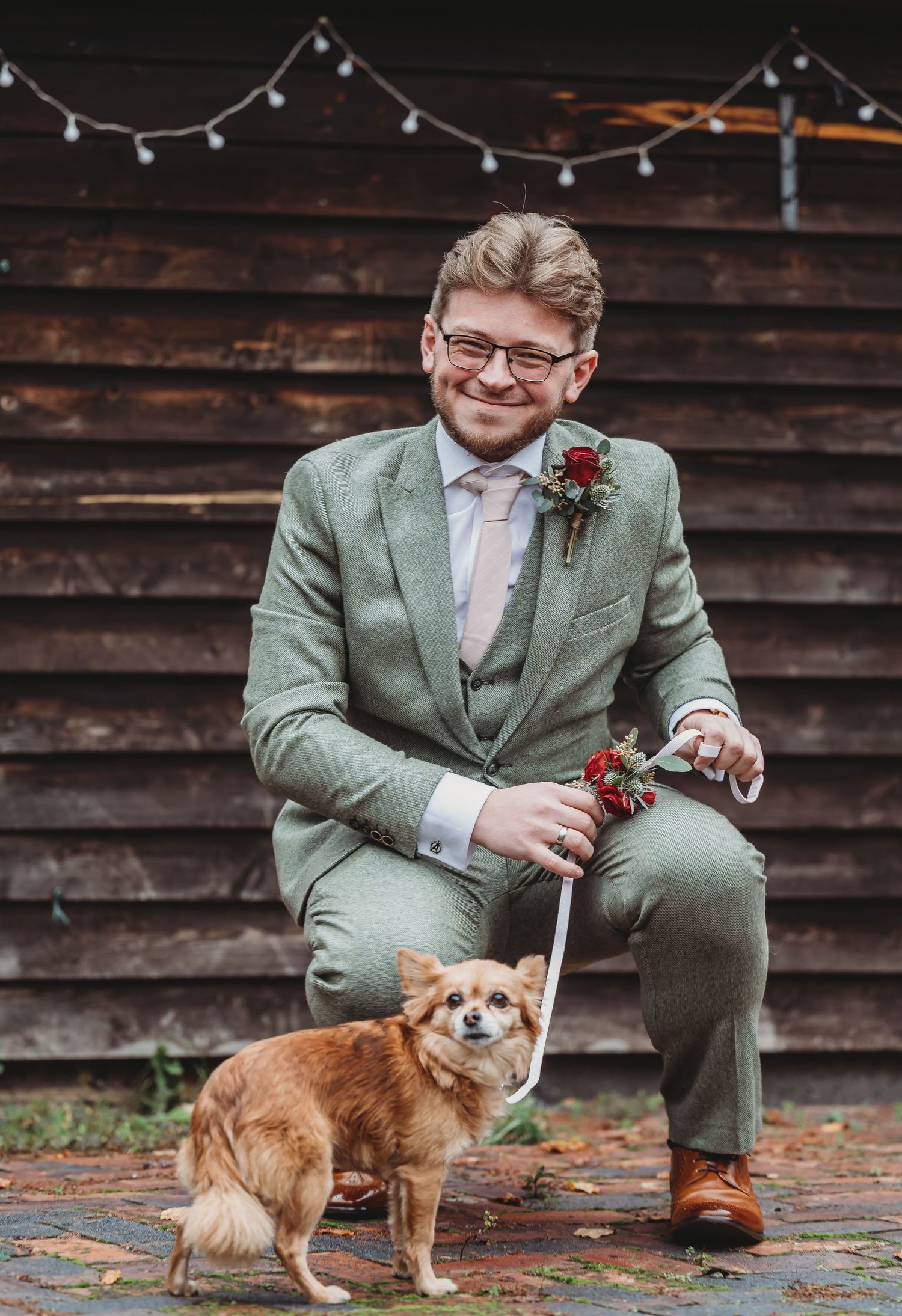 A dog accompanies someone wearing a sage green suit at a rustic wedding venue with string lights.
