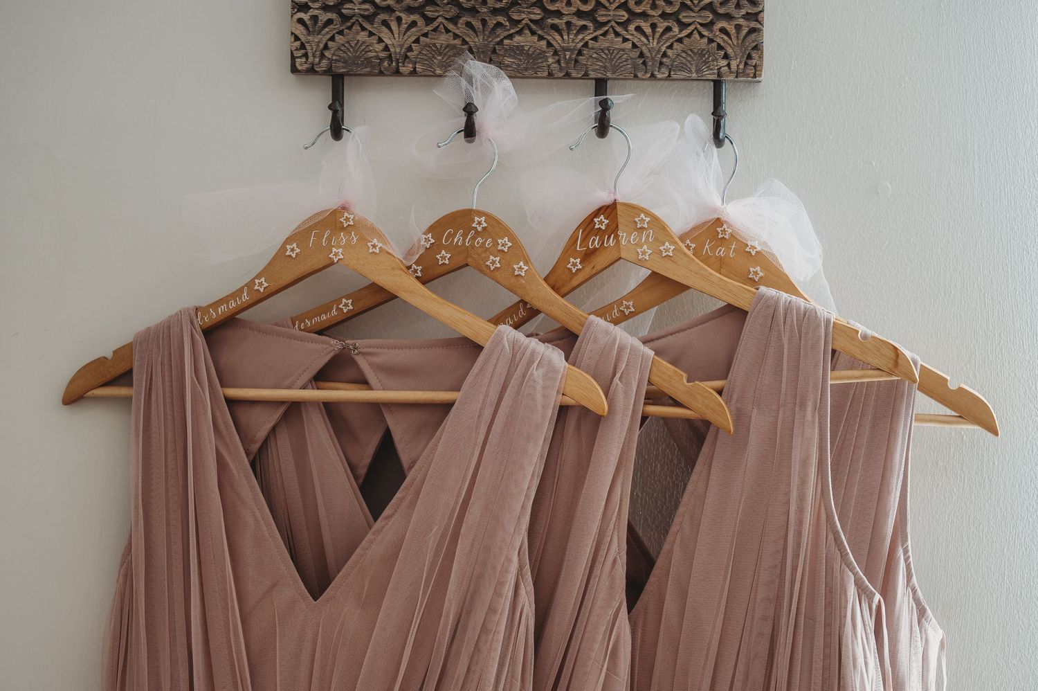 Blush pink bridesmaid dresses hanging on wooden hangers against a decorative wall.
