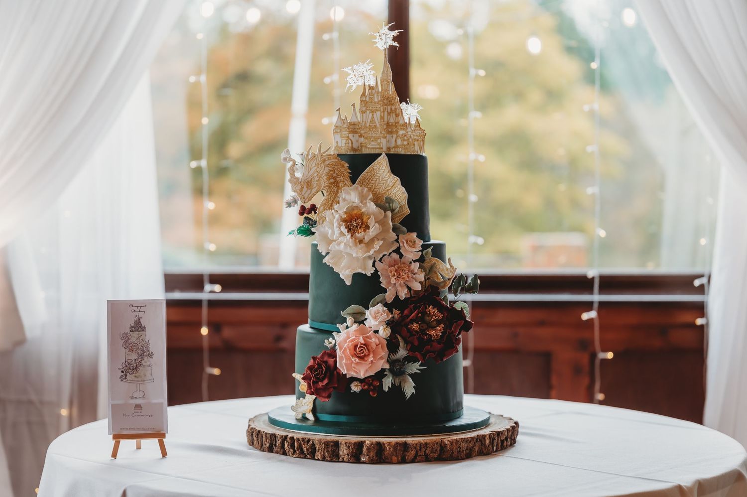 Elegant dark green and gold wedding cake with peach flowers displayed on a wooden cake stand.