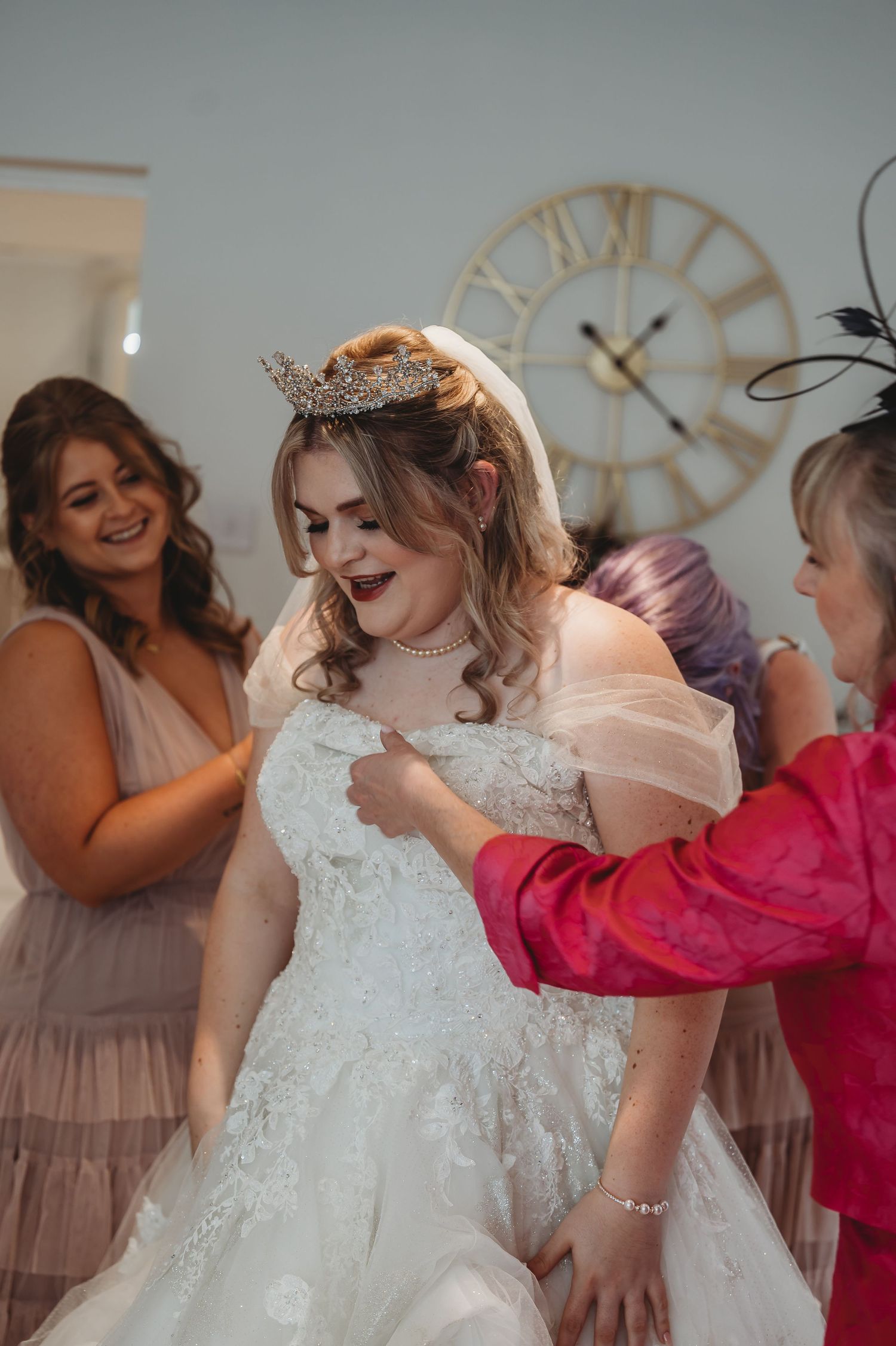 Wedding preparations show helpers assisting with a bride's white lace gown under a decorative wall clock.