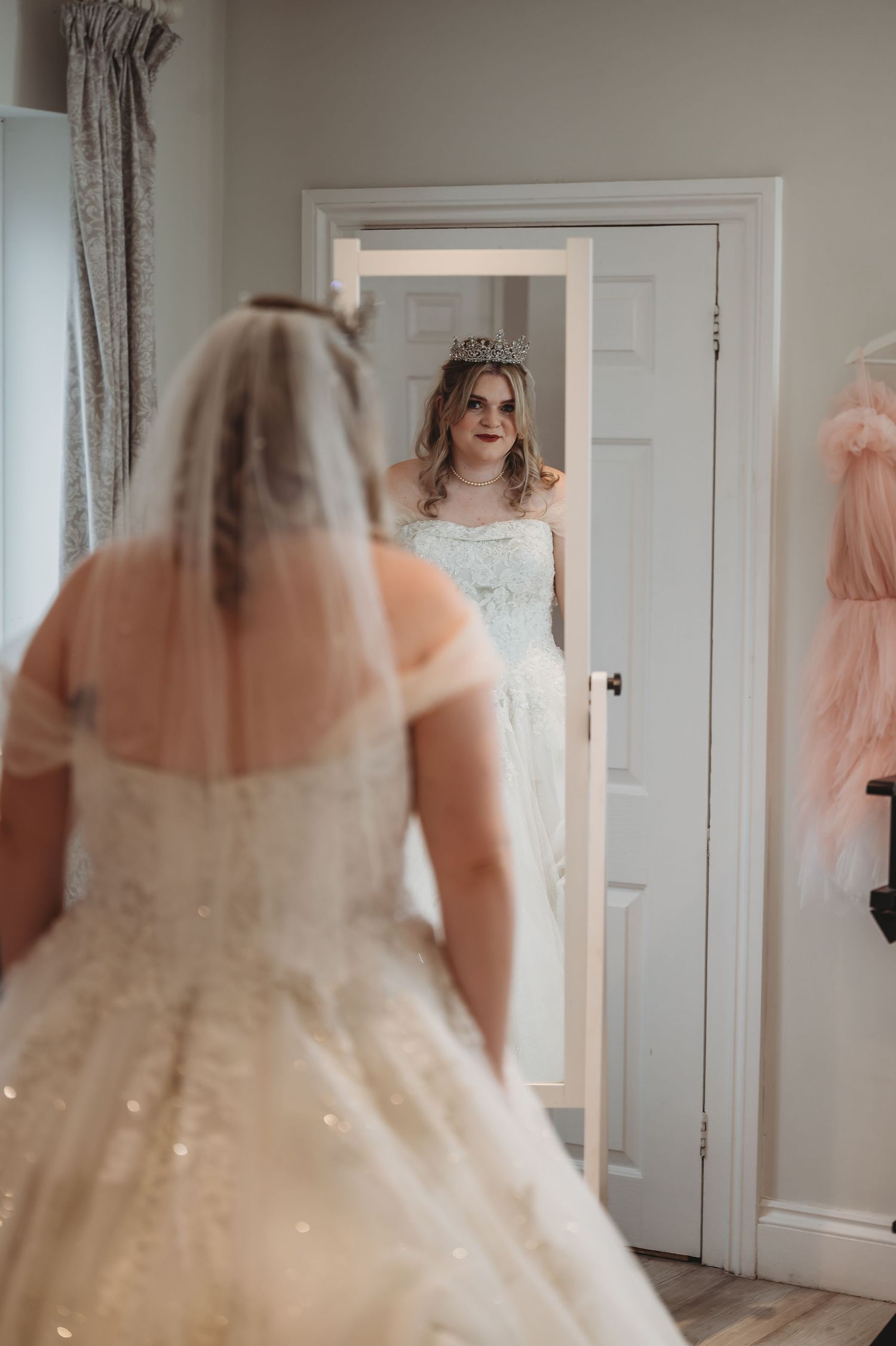 A bride in a sparkly ball gown admires her reflection in a full-length mirror while wearing a tiara.
