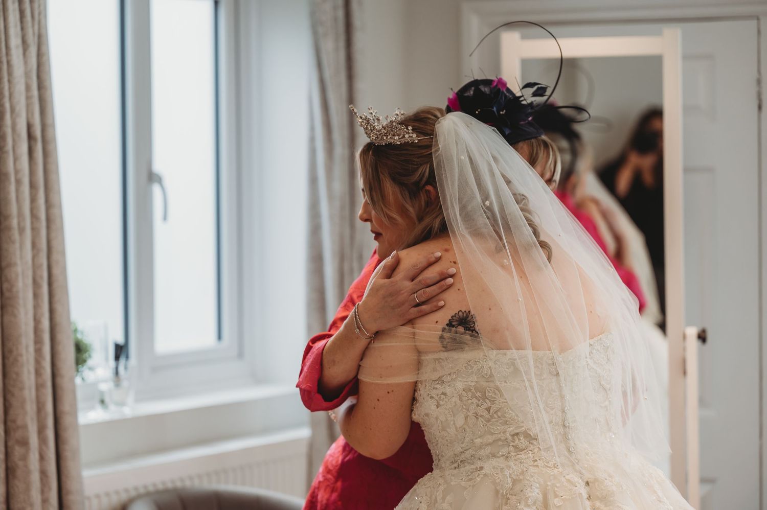 An emotional embrace between two people wearing bridal attire and red clothing in a sunlit room.