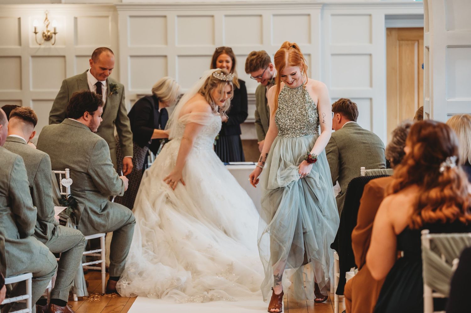 Wedding guests gather in an elegant venue with white paneled walls as a bride twirls in her flowing gown.