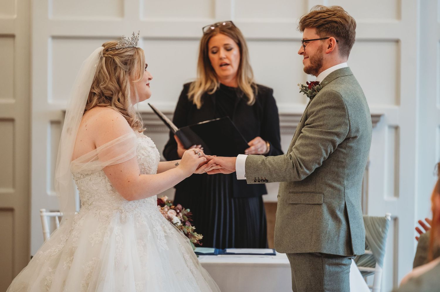 A wedding ceremony unfolds in a bright interior with white wall panels as vows are exchanged.