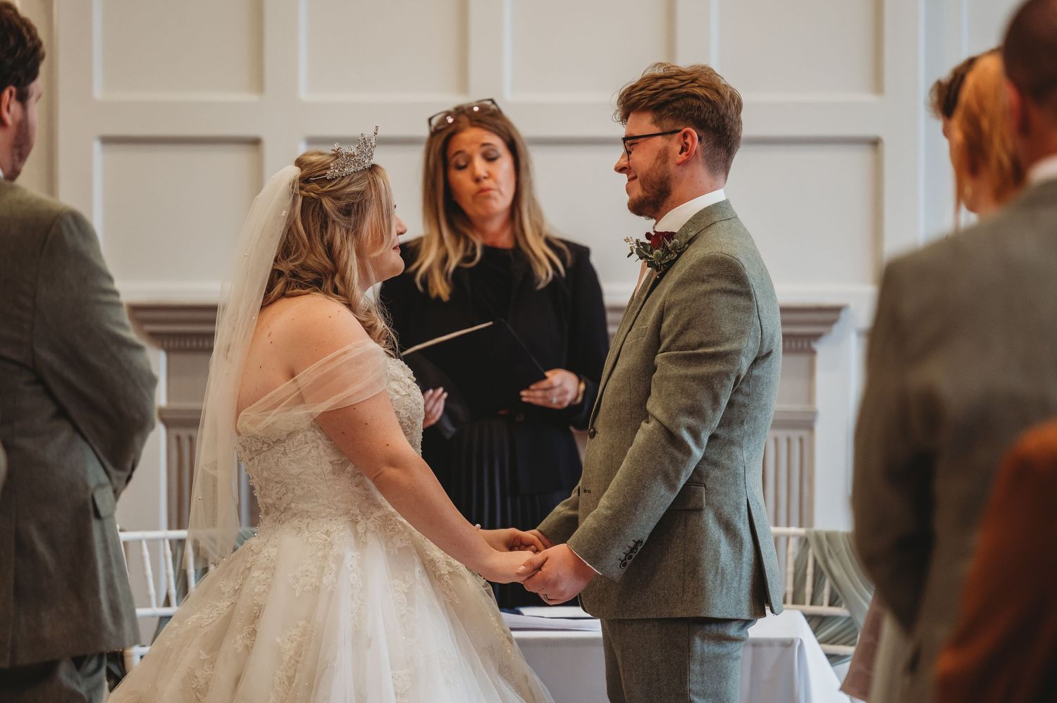 A wedding ceremony takes place in a bright room with white paneled walls as a couple exchanges vows before an officiant.