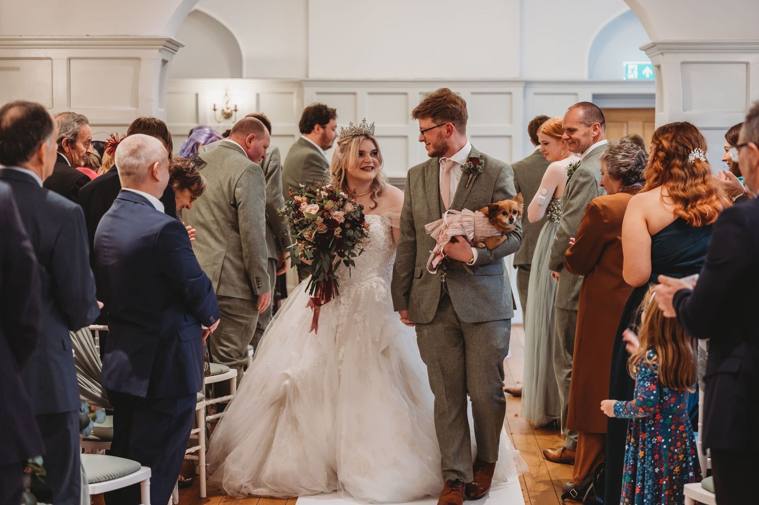 Happy couple share a moment walking down the aisle surrounded by wedding guests and white decor.
